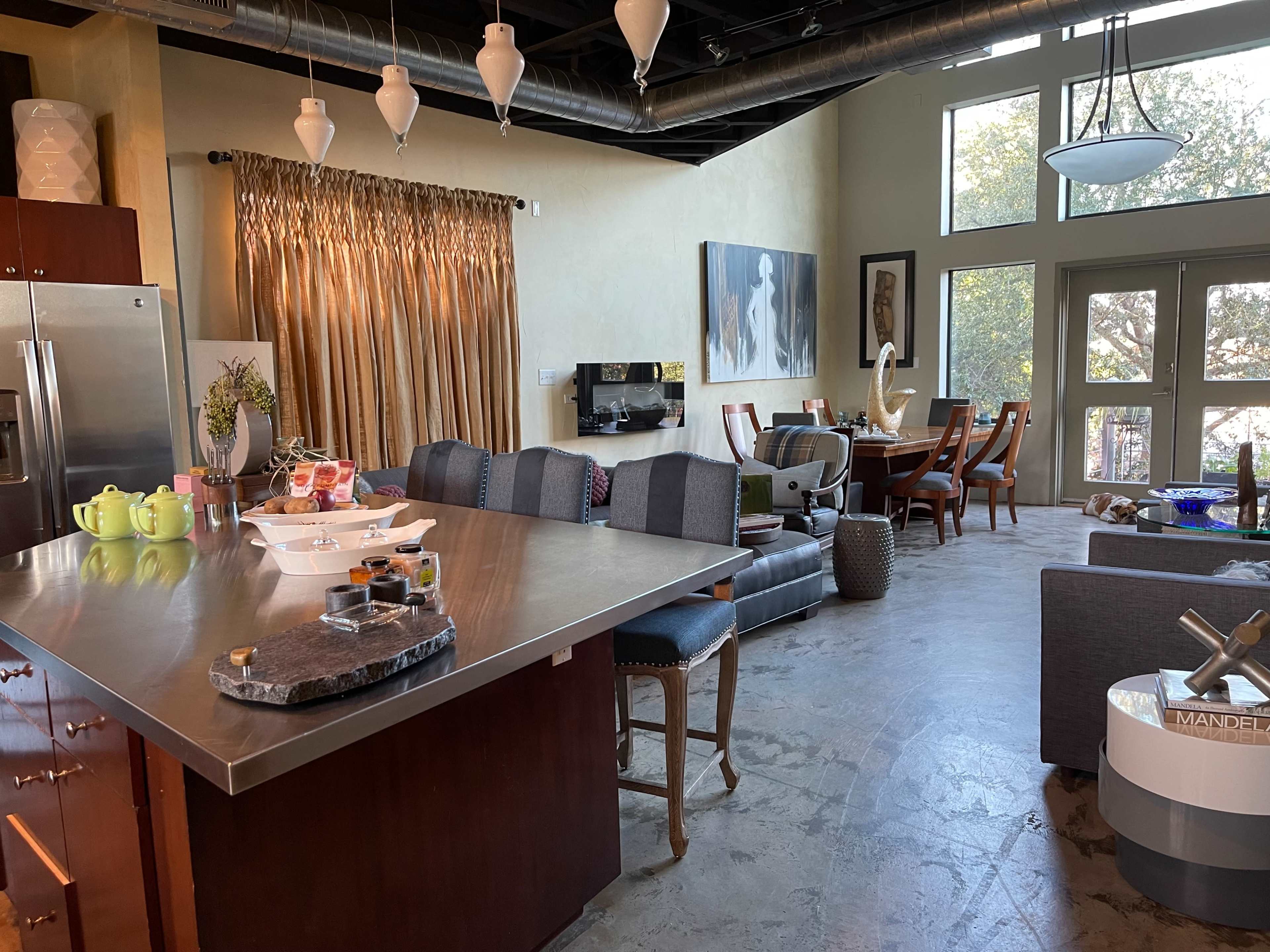 The image shows a modern kitchen and living area with a large island, dining table, and windows letting in natural light.