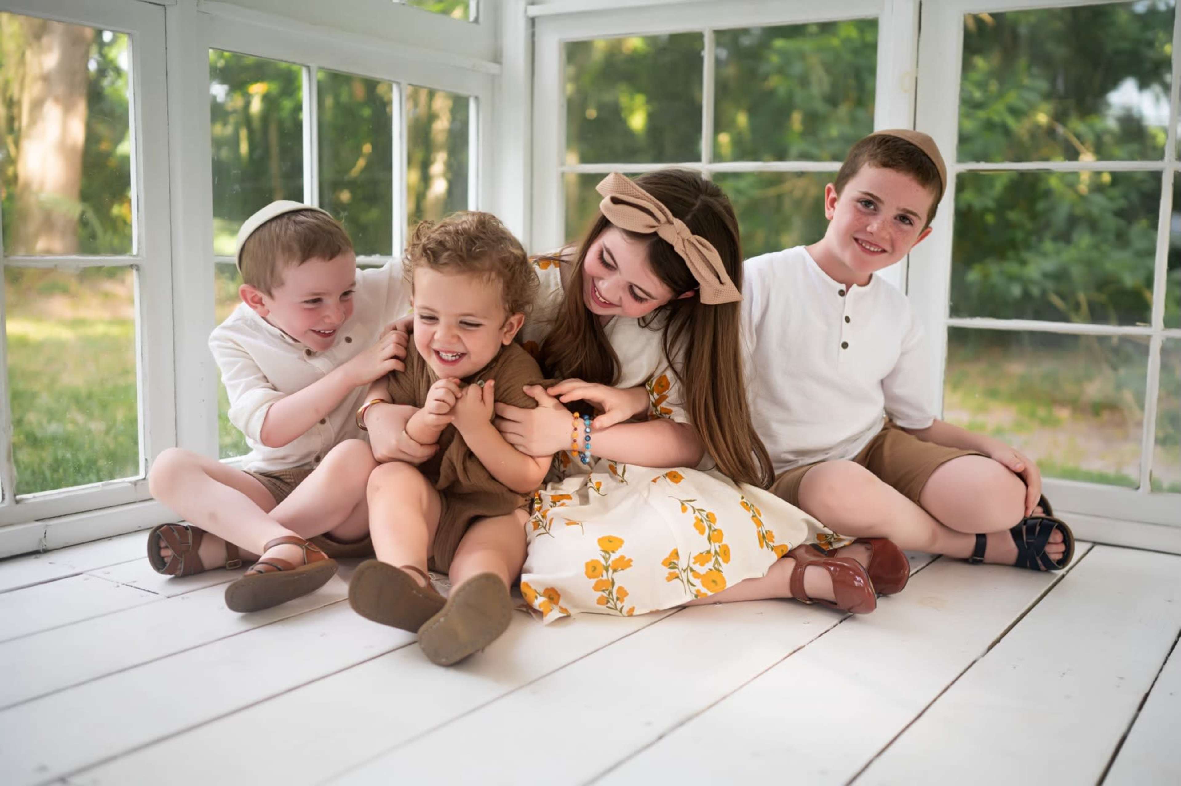 Four children are sitting on a white wooden floor in a sunlit room with large windows, playfully interacting with each other.