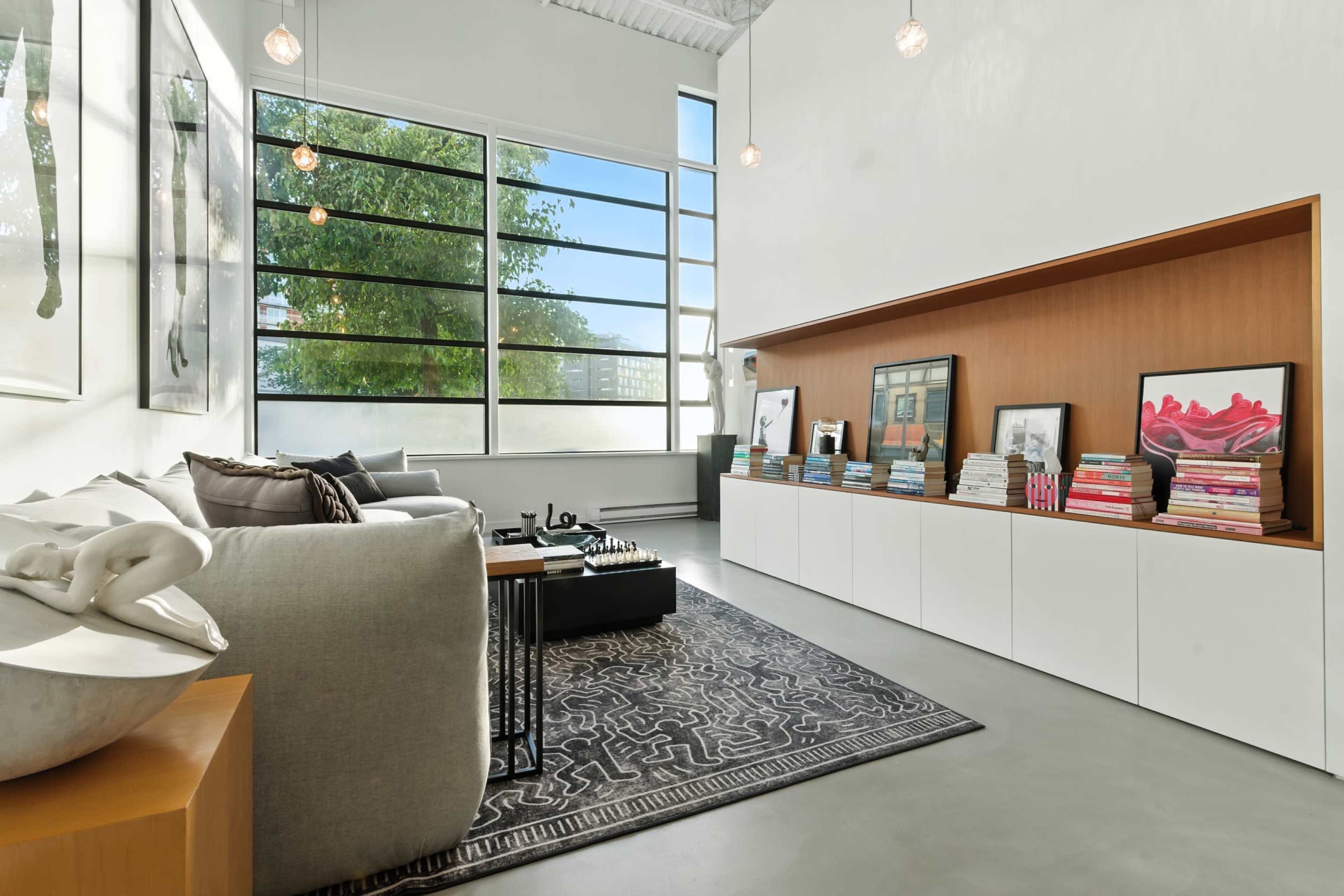 A modern living room features large windows, a minimalist design with a gray sofa, a wooden storage unit, and a dark area rug.
