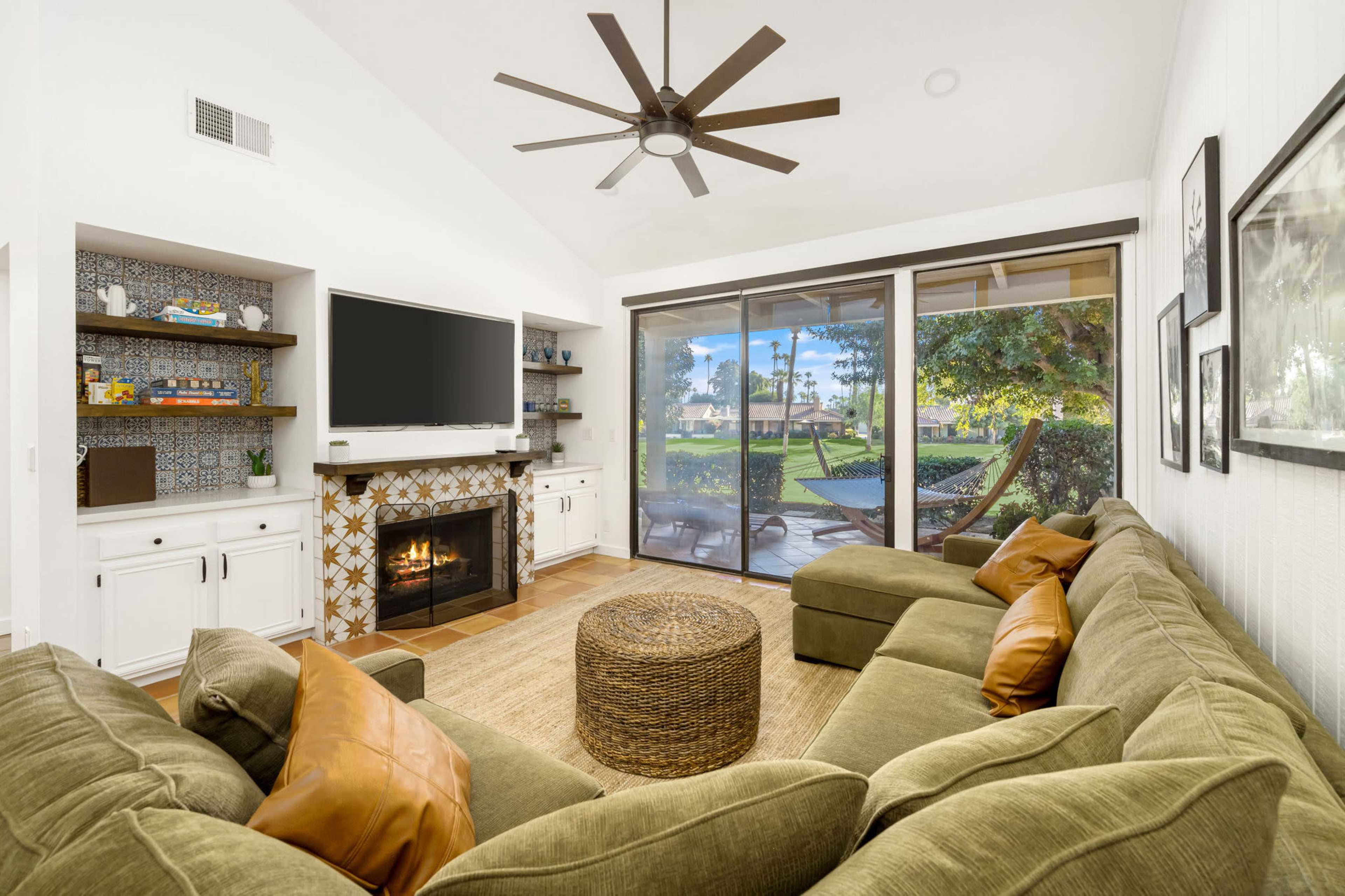 A cozy living room features a large sectional sofa, a mounted television, and a fireplace, with sliding glass doors opening to a view of the outdoor area.