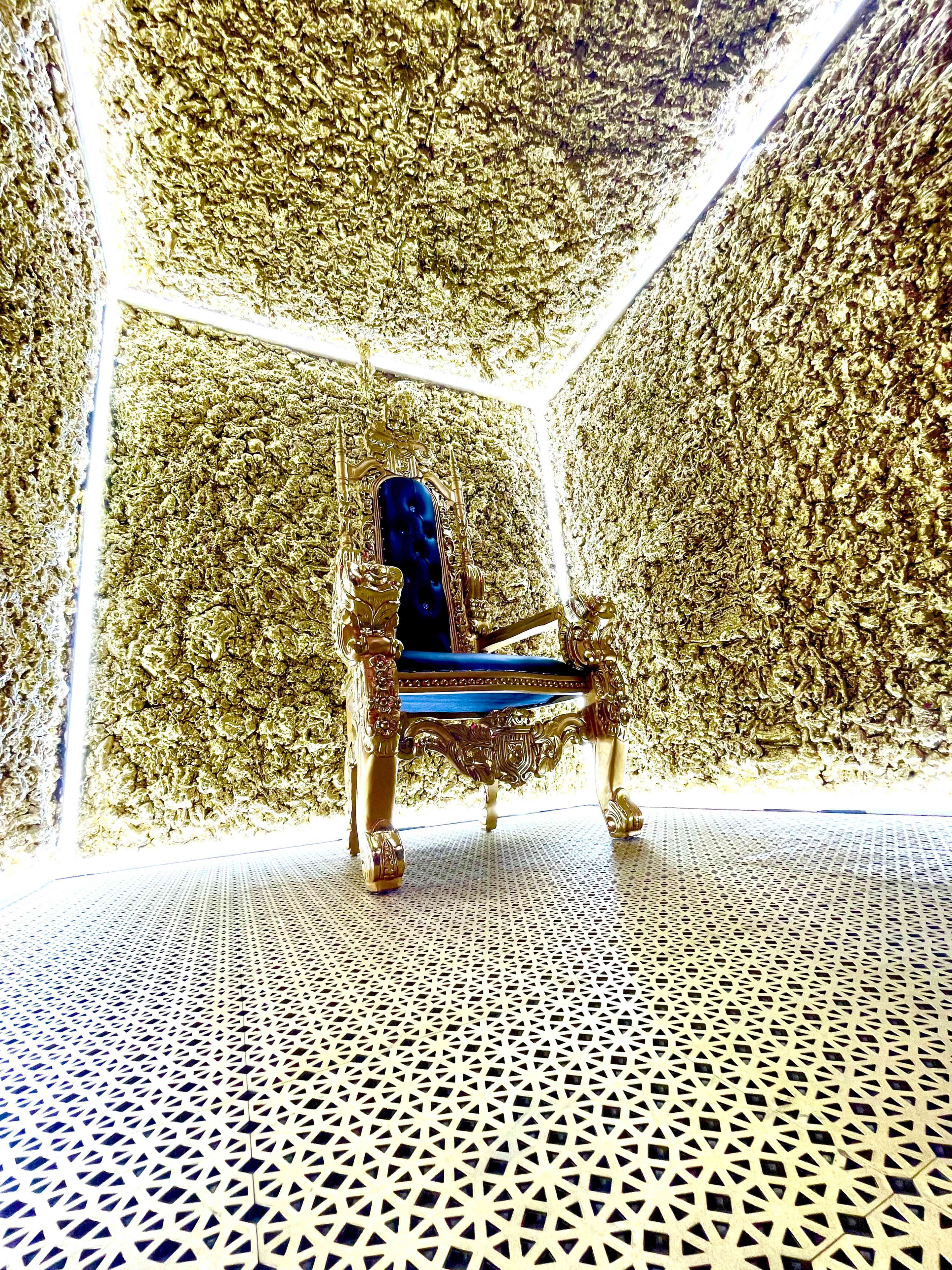An ornate golden throne placed against walls covered in textured, golden material, illuminated by bright light from above.