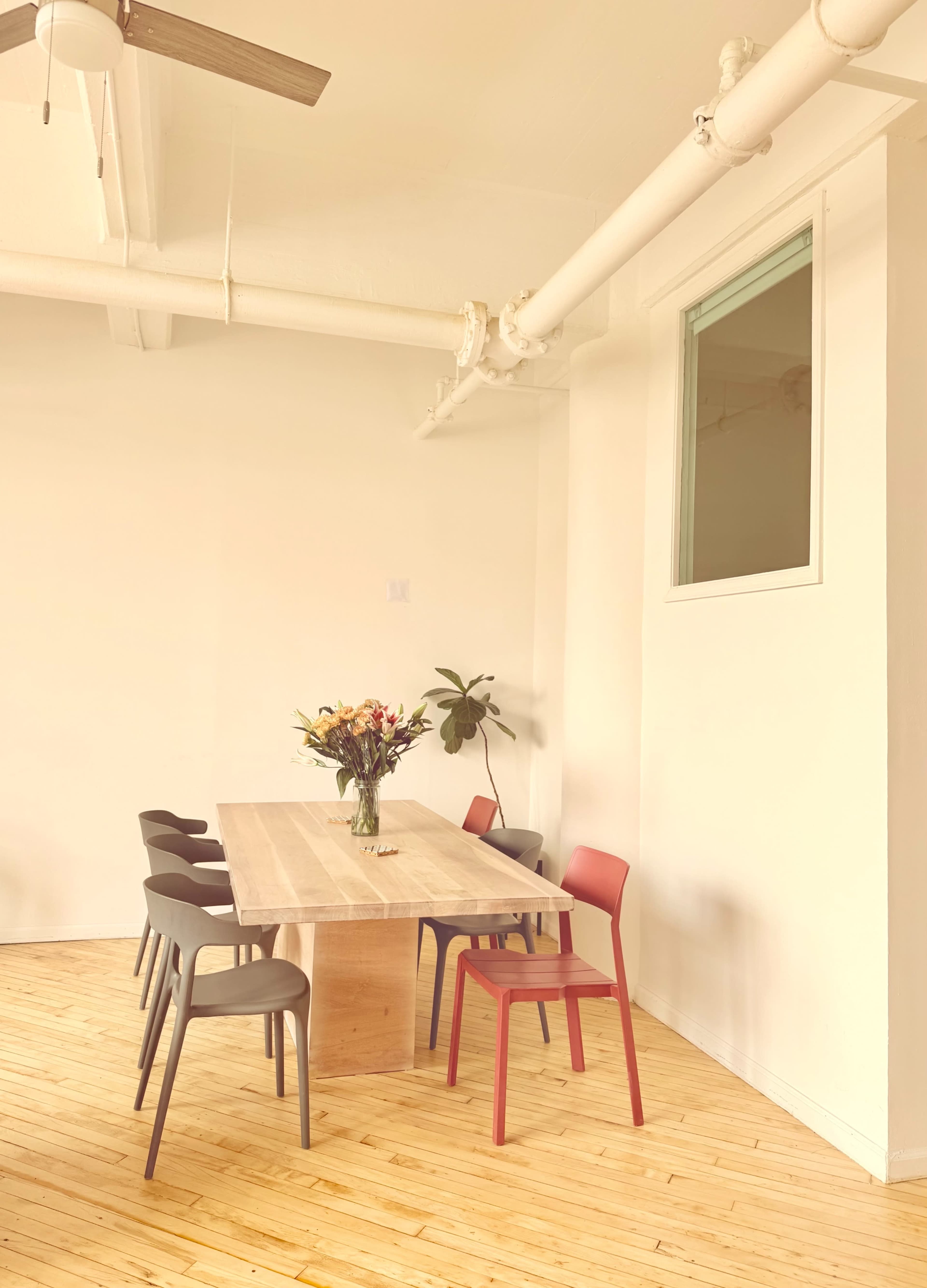 A wooden table with a vase of flowers is surrounded by six gray chairs and one red chair in a brightly lit room with wooden flooring.