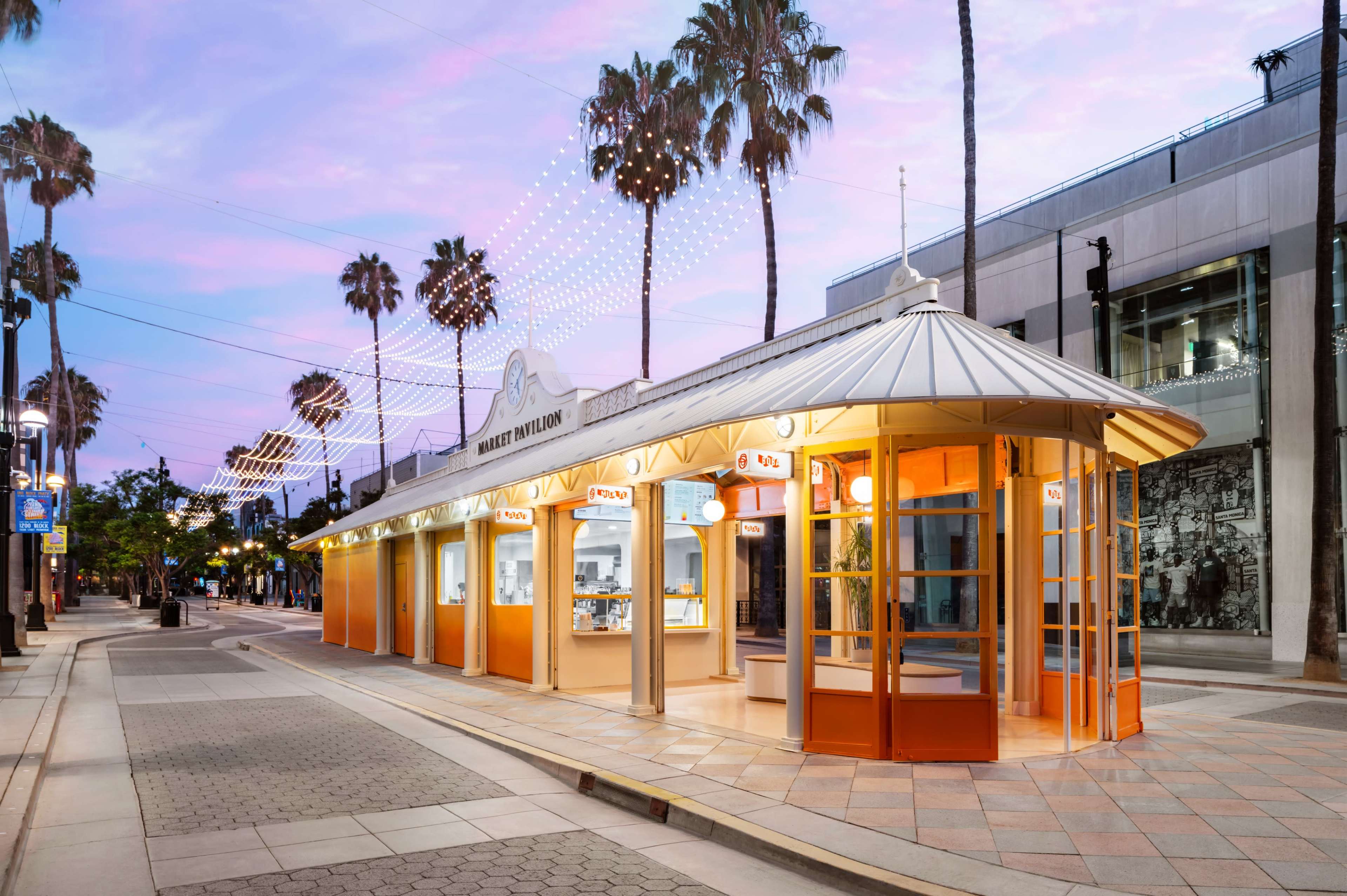 The image shows a brightly lit pavilion with an orange and white exterior, situated along a palm tree-lined street at dusk.