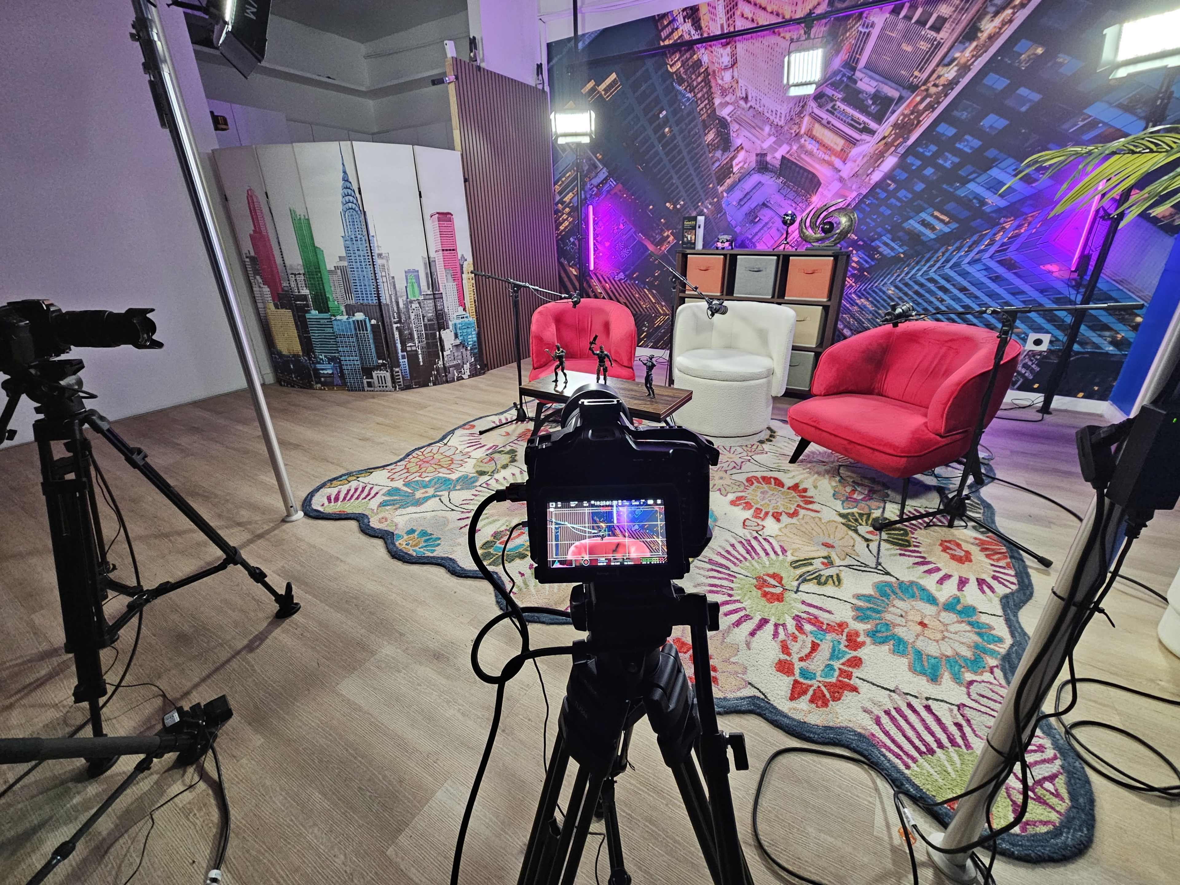 The image shows a brightly lit studio setup for a video recording, featuring pink chairs, a colorful rug, and cameras arranged to capture the scene against a backdrop of a cityscape.