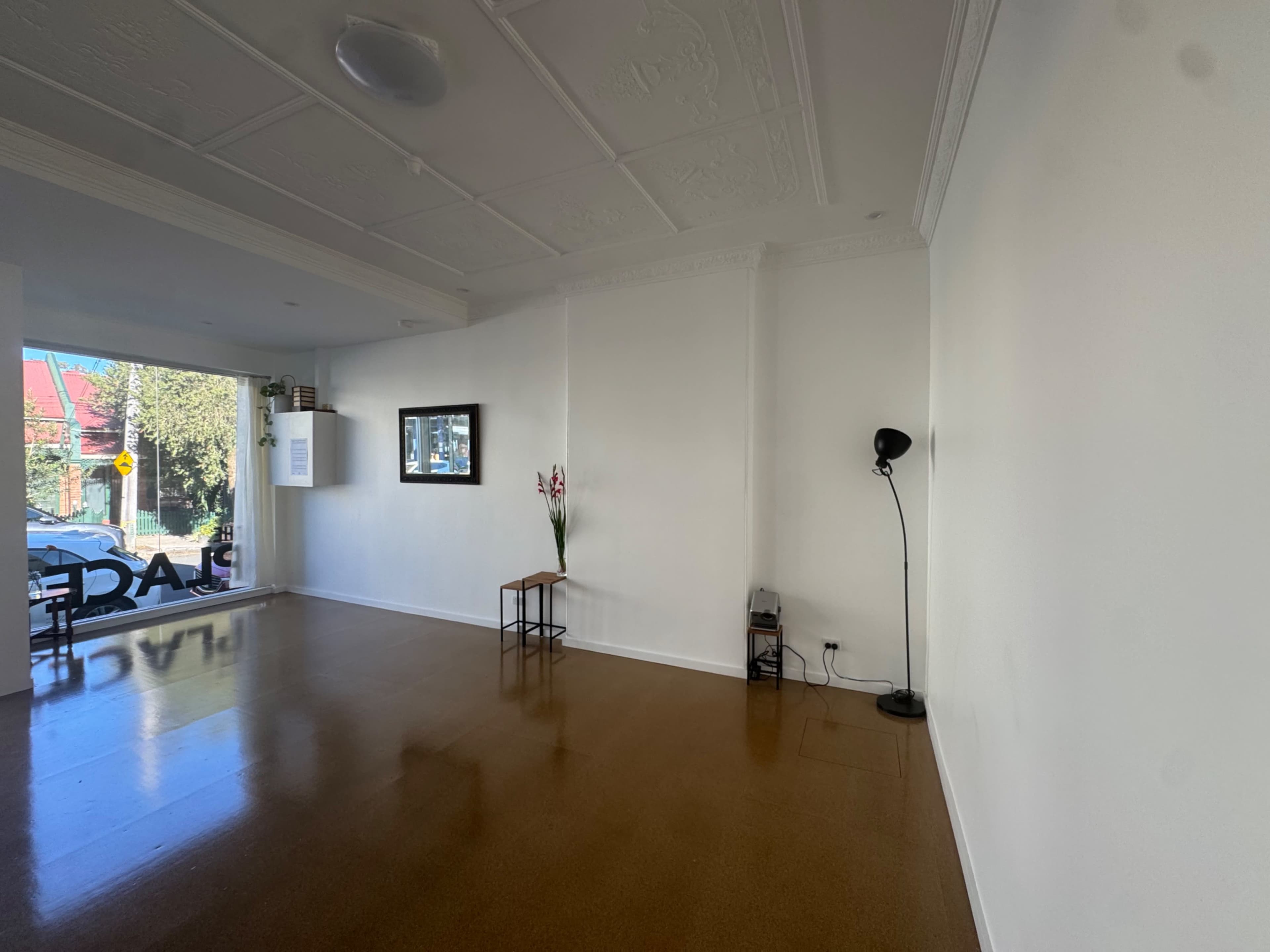 The image shows an empty room with a glossy floor, a small table, a floor lamp, and a window displaying a view of the street outside.
