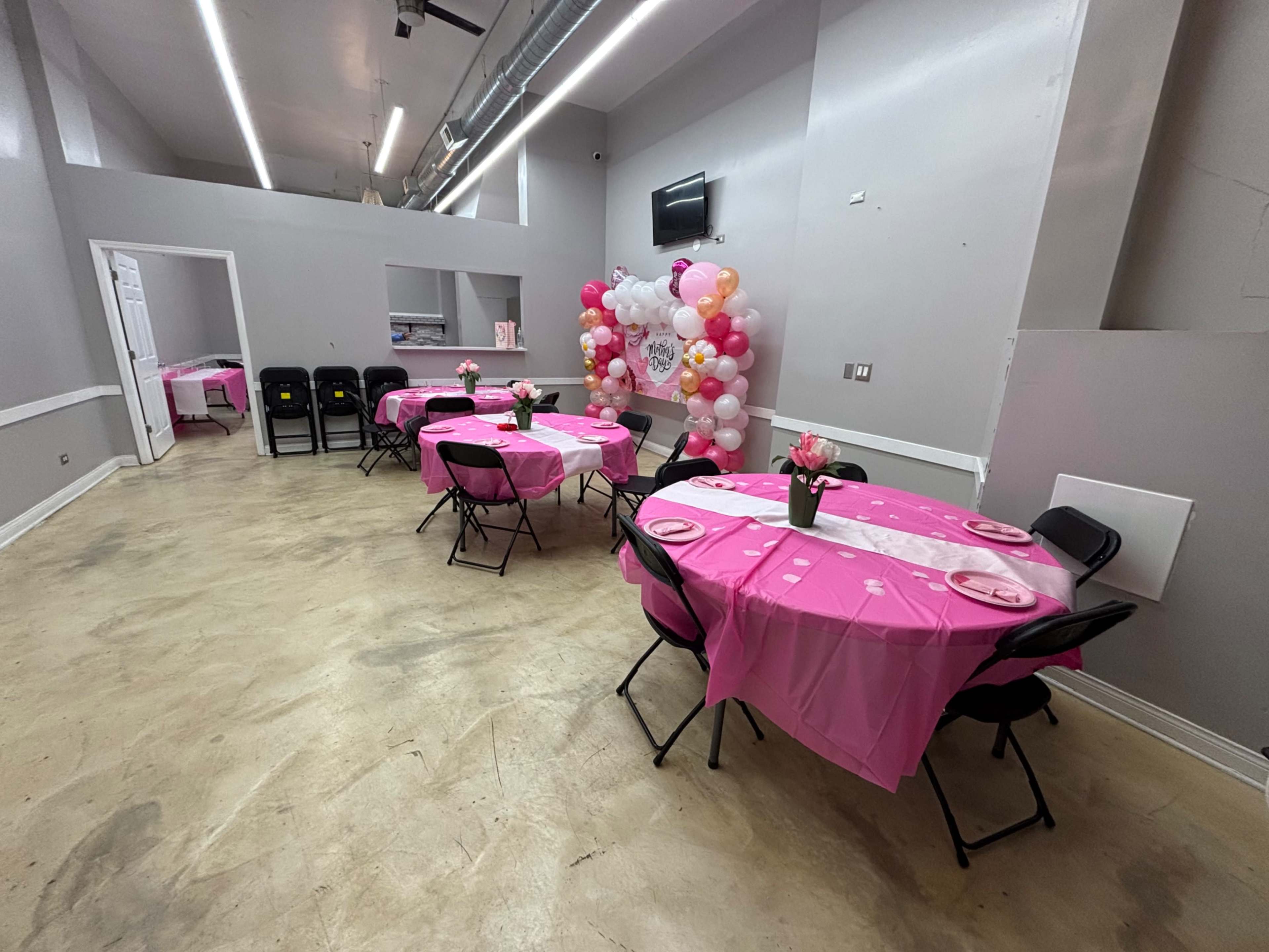 The image shows a party venue set up with round tables covered in pink tablecloths, black chairs, and a decorative balloon display in the background.