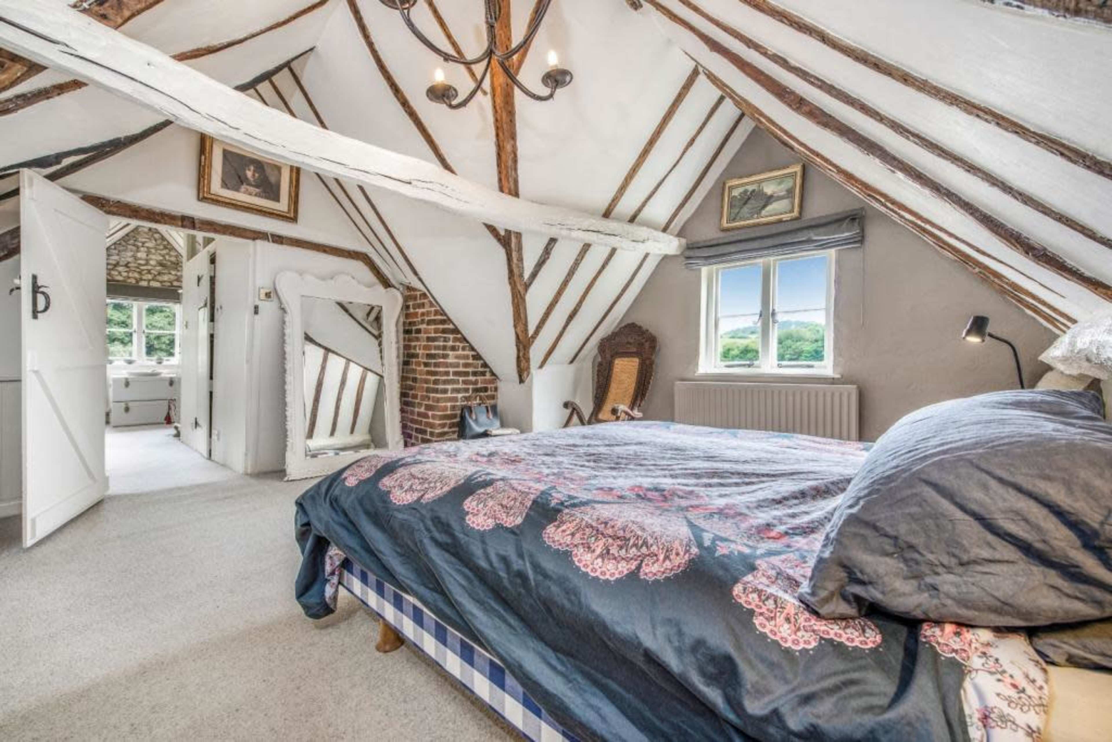 The image shows a cozy attic bedroom with exposed wooden beams, a large bed covered in a dark quilt, and an adjacent doorway leading to a bathroom.