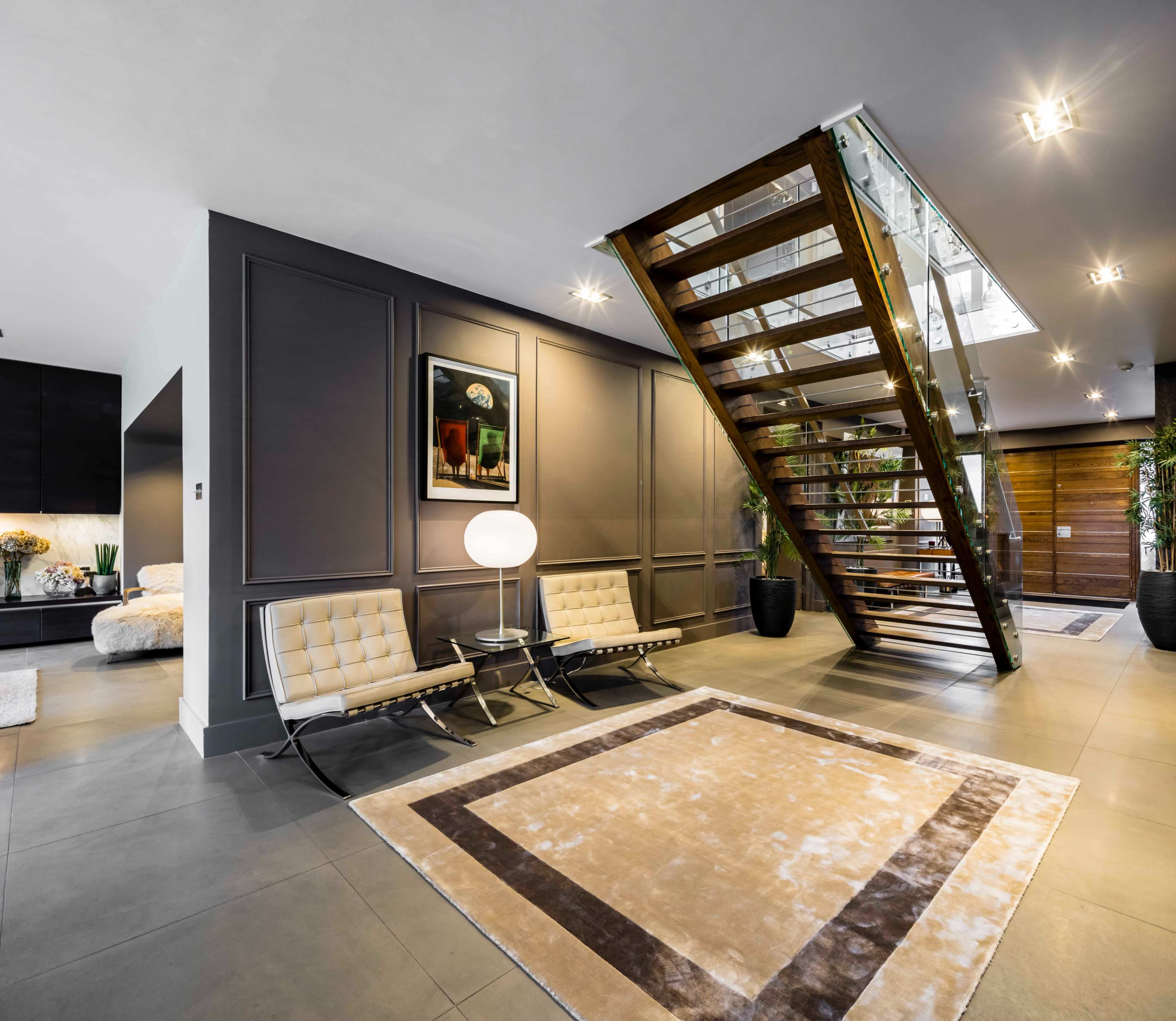 A modern foyer features a wooden staircase, sleek seating, a round lamp, and a large area rug with a geometric design.
