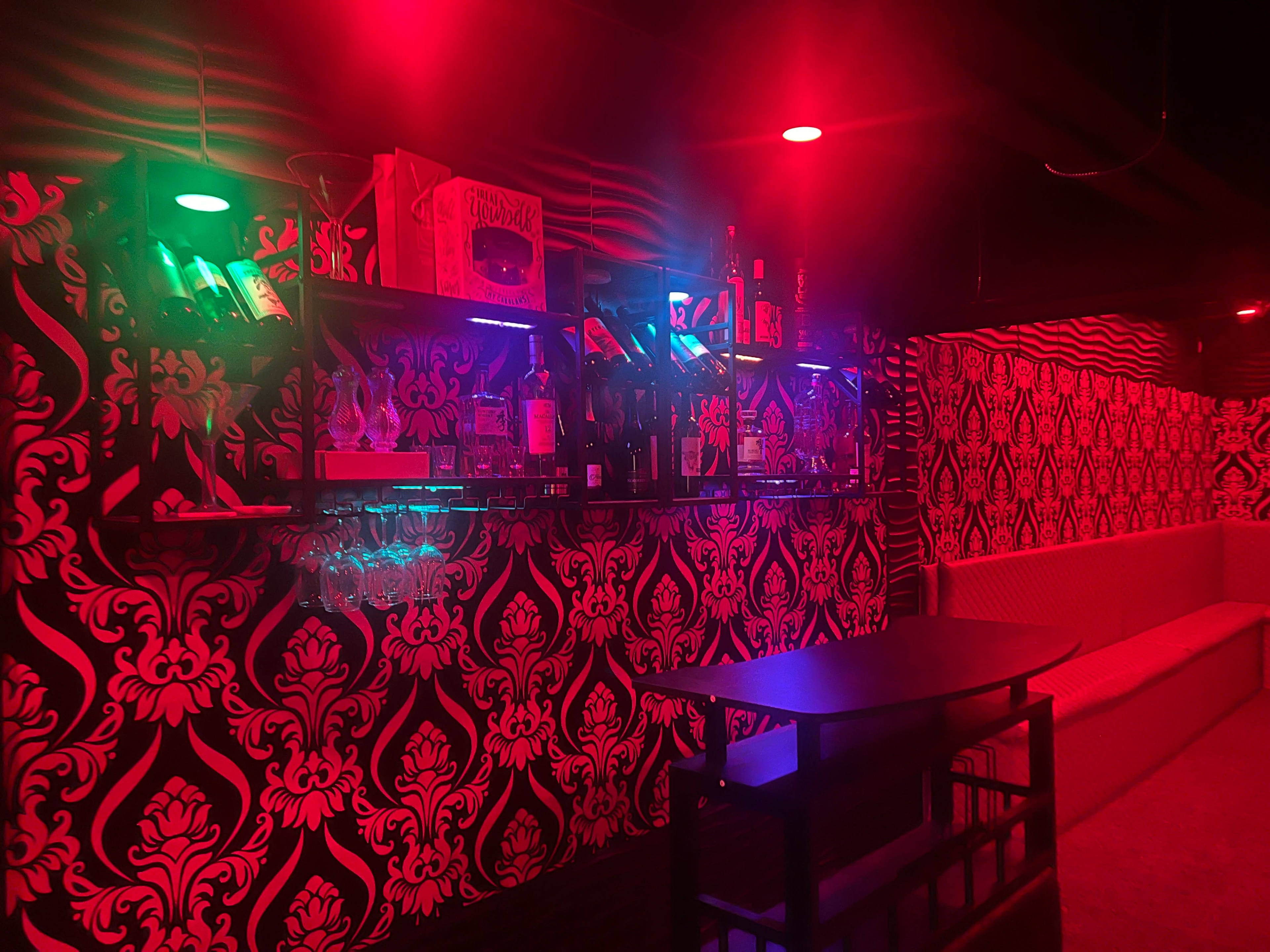 The image shows a dimly lit bar area with a patterned black and red wall, illuminated by colorful lights, featuring shelves stocked with bottles and glasses.