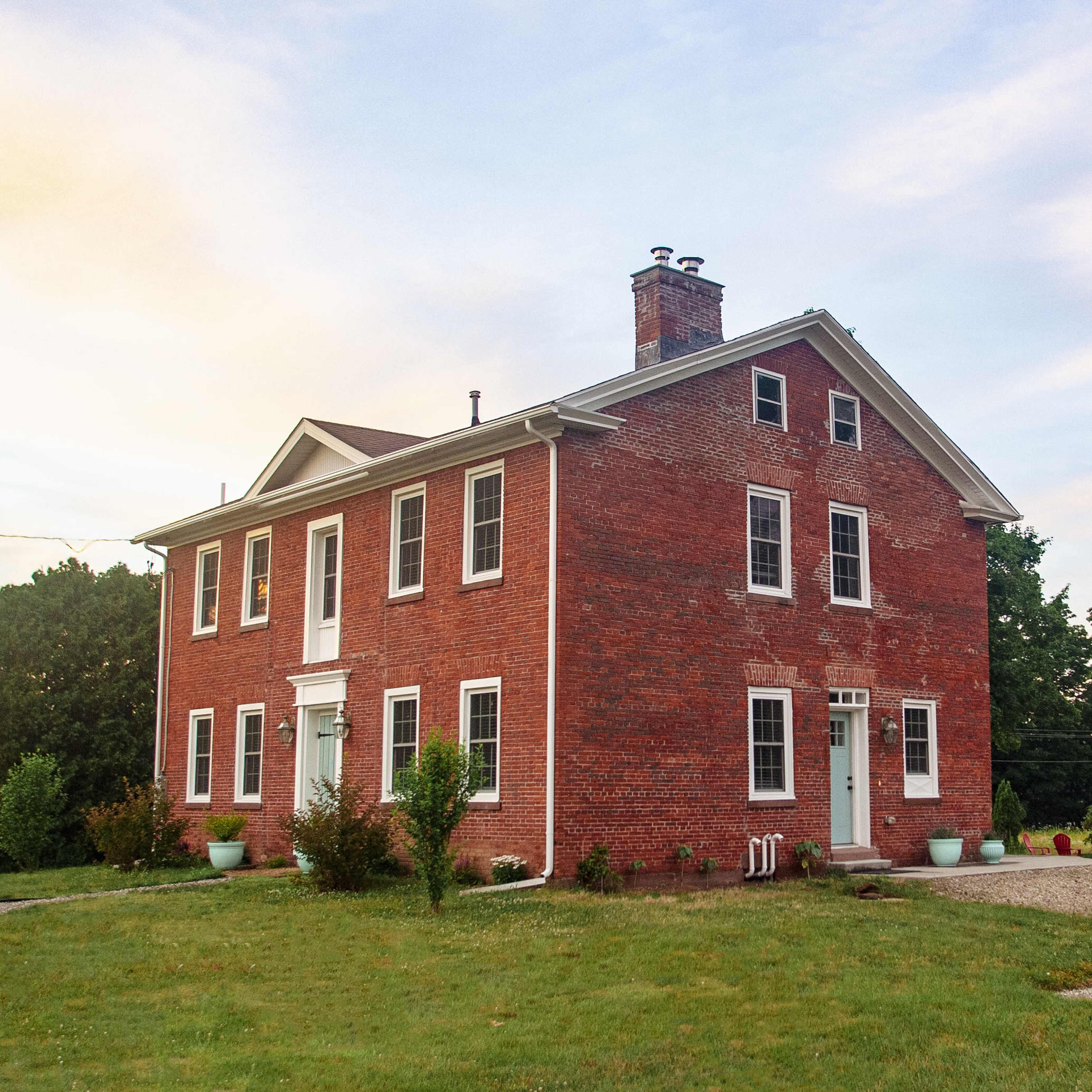 A two-story red brick house with white trim, a central front door, and several windows sits on a grassy lot.