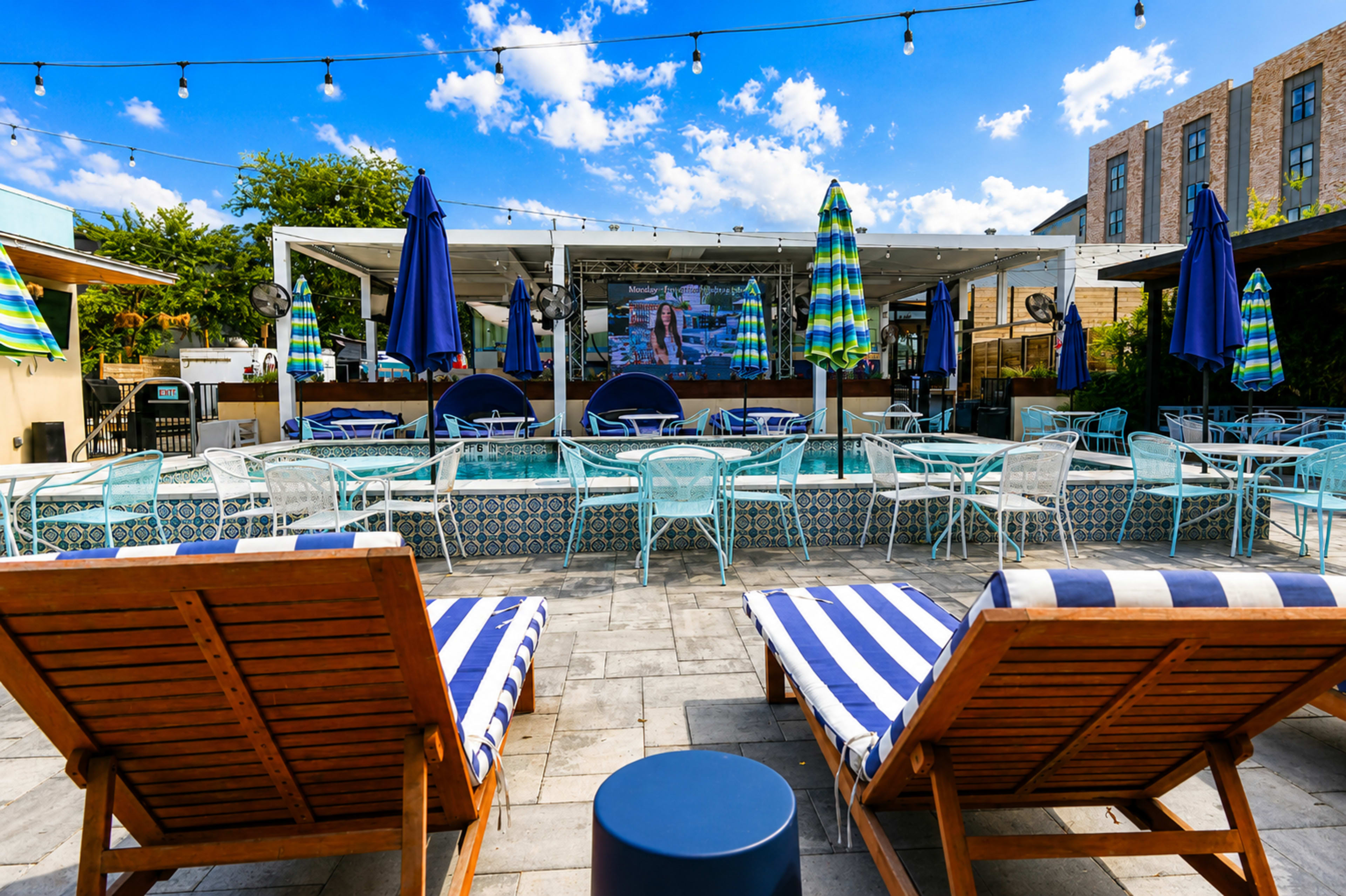 The image shows a poolside area with striped lounge chairs, outdoor seating, and a large screen, all set against a bright blue sky with scattered clouds.