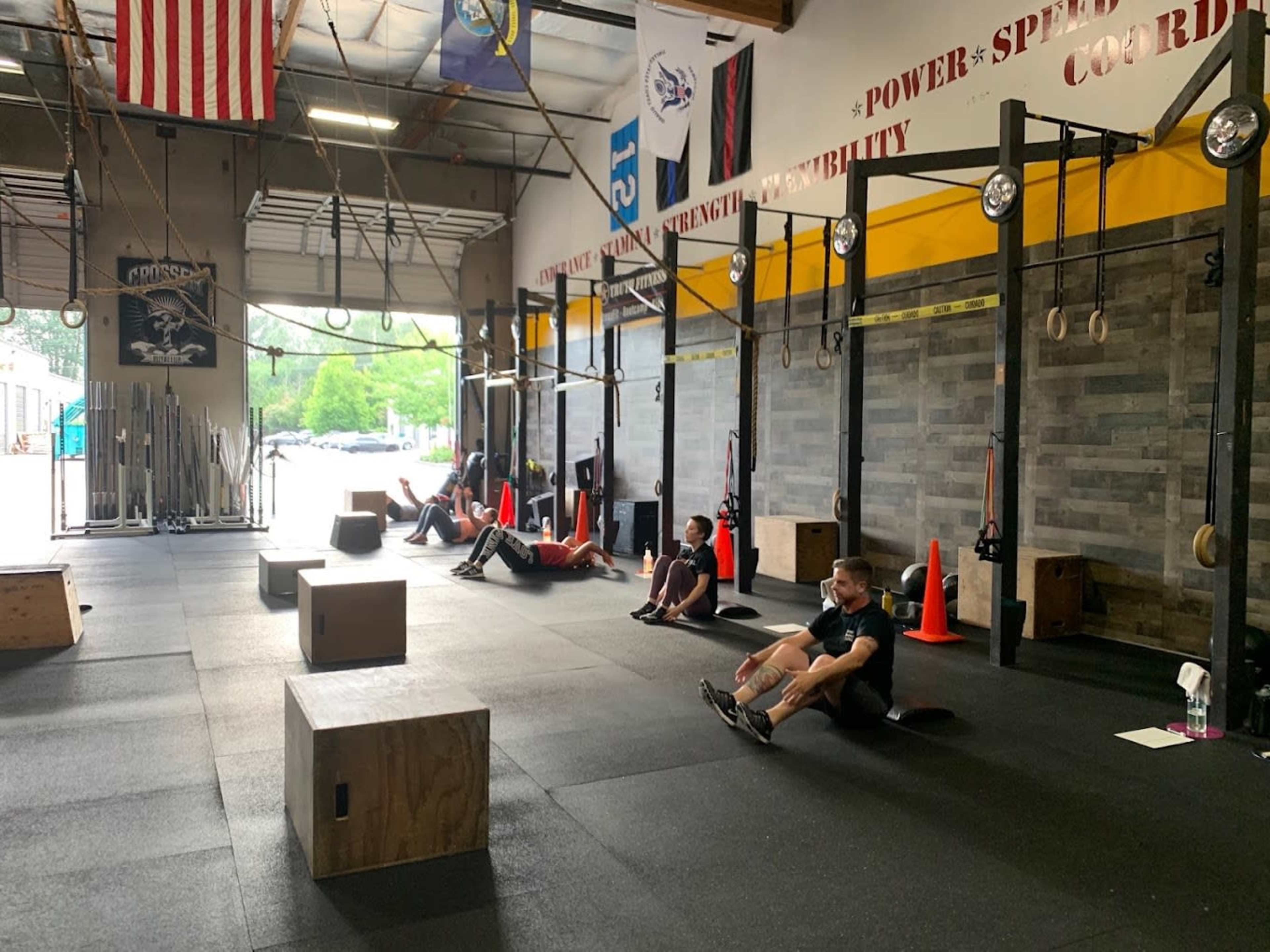 A group of individuals is seated on the floor of a gym, with exercise equipment and boxes in the background.