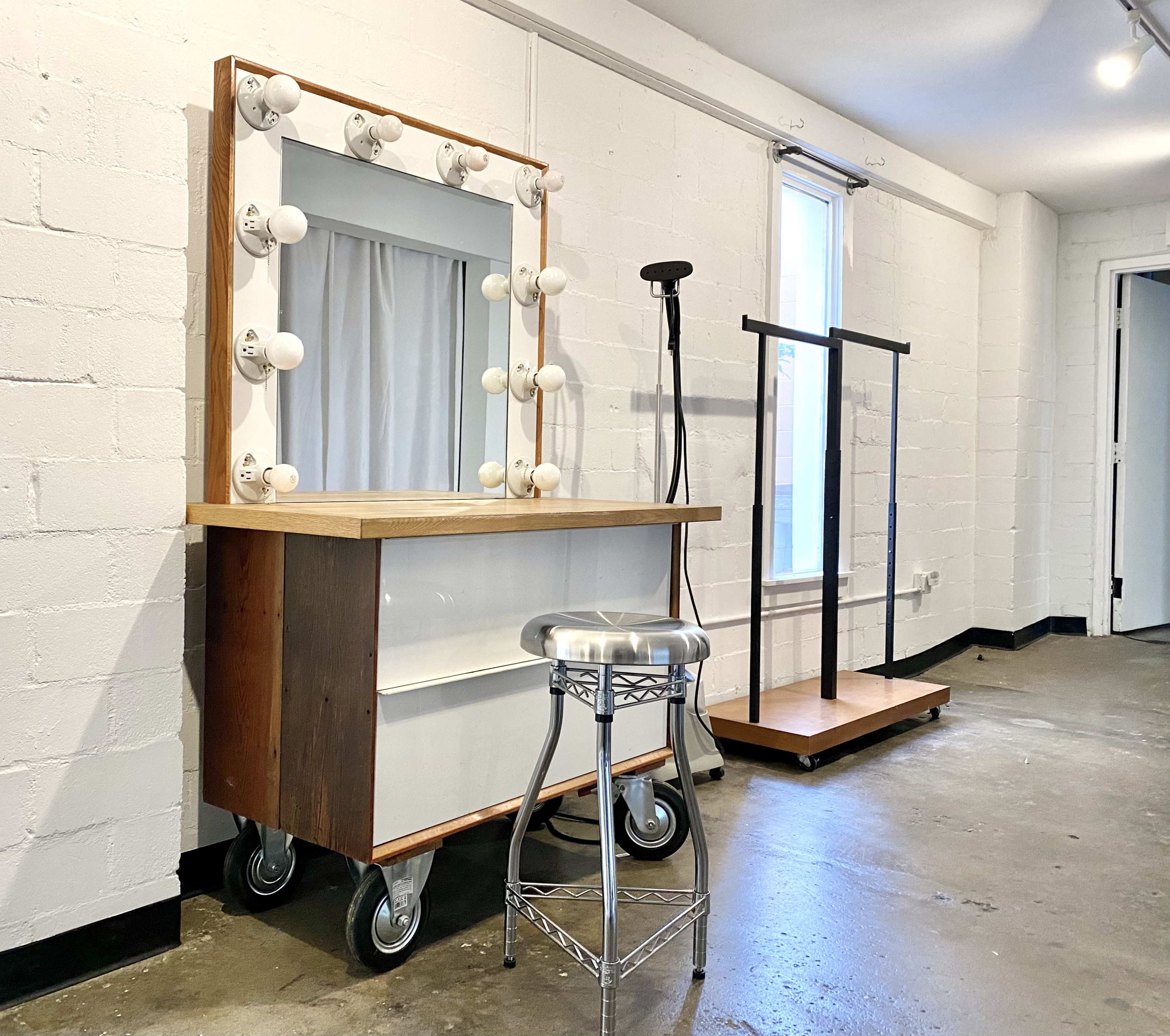 A makeup vanity with a large illuminated mirror, a metal stool, and a clothing rack in a minimalist room with white walls and concrete flooring.