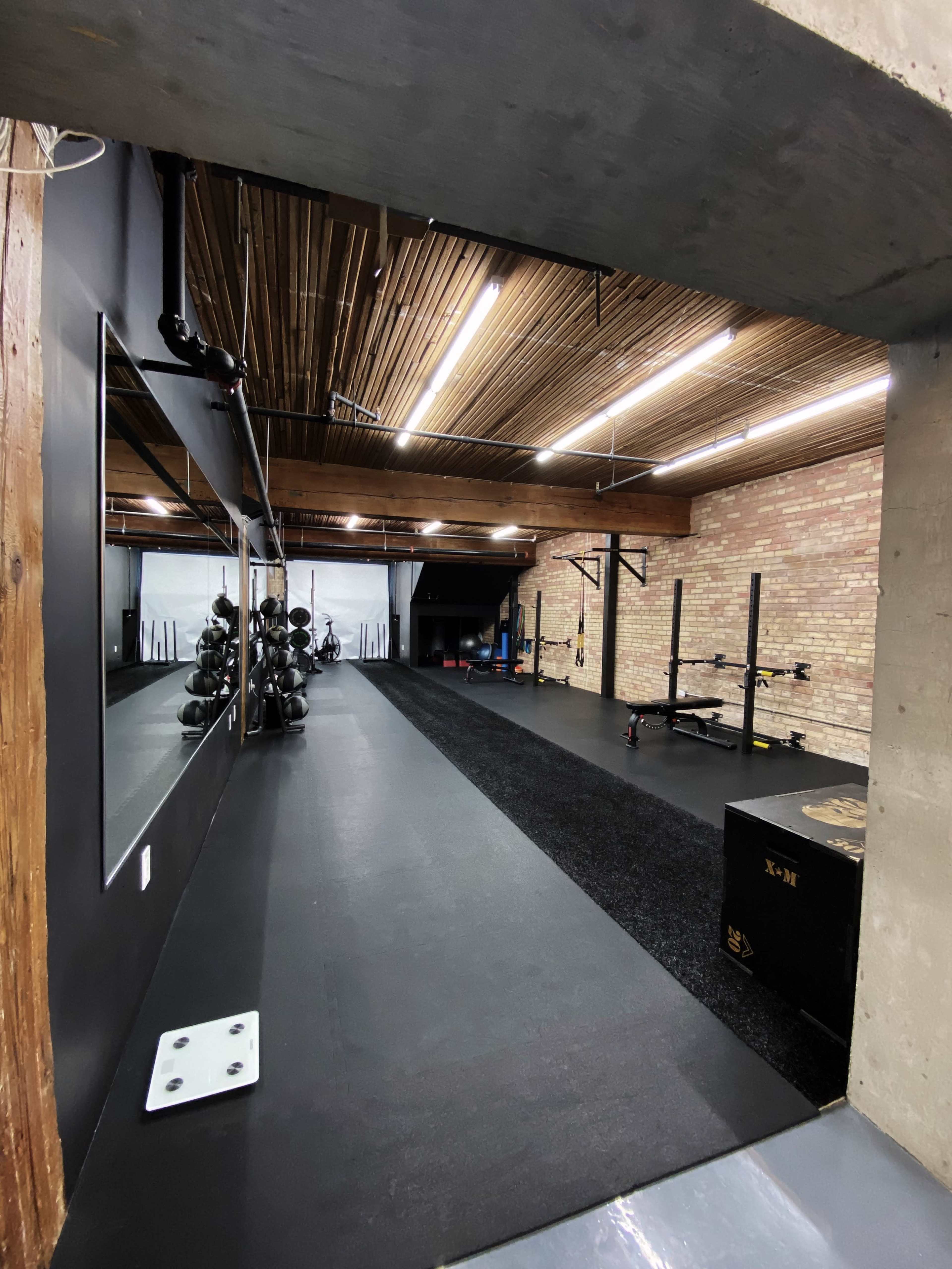 A spacious, modern gym with black rubber flooring, exposed brick walls, and various exercise equipment along the mirrored wall.