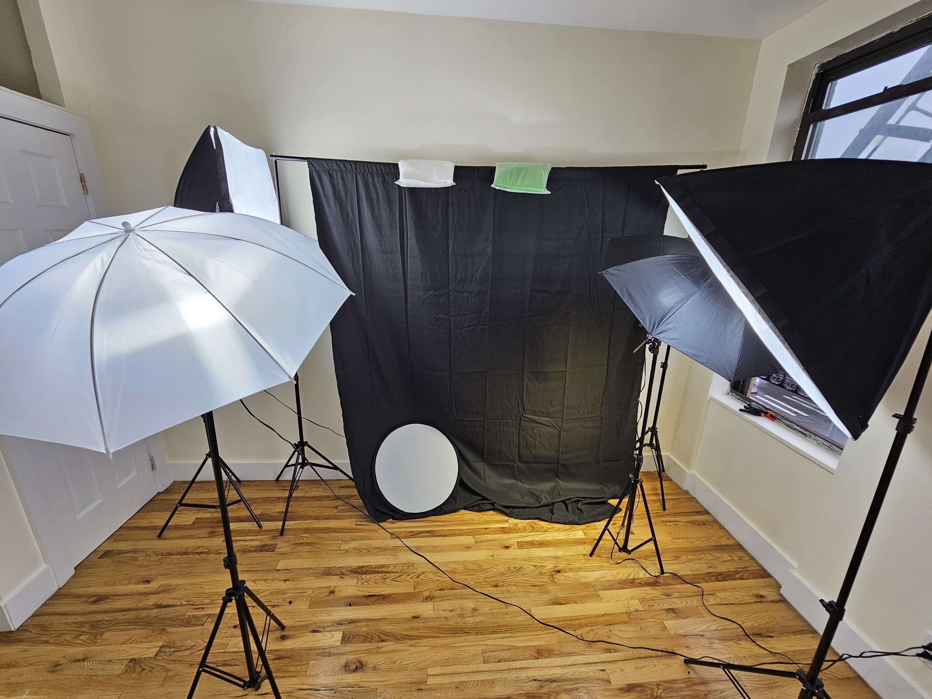 A photography setup with two softbox lights and an umbrella light positioned around a black backdrop in a room with wooden flooring.