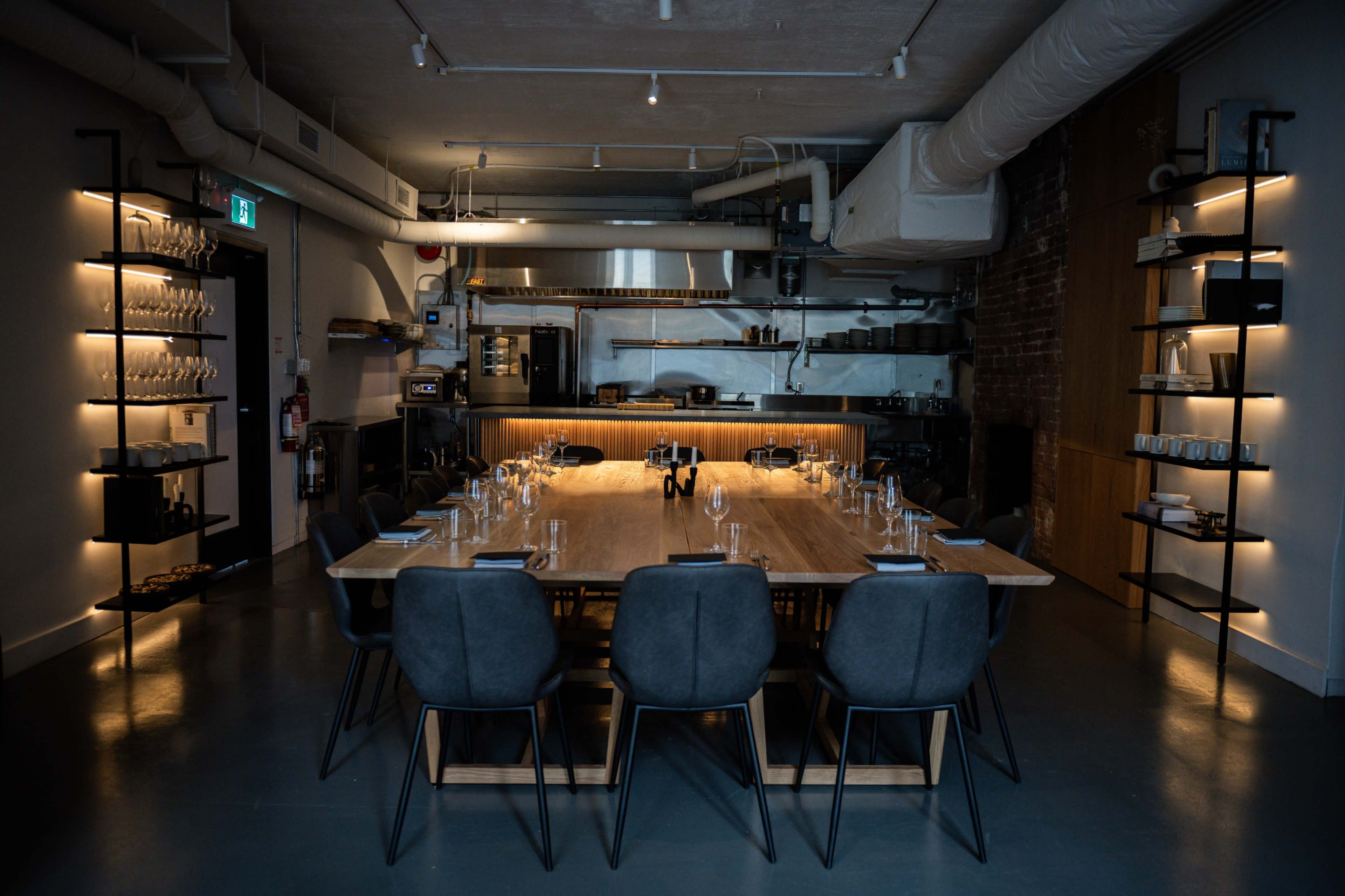 A long wooden dining table is set with glasses and plates in a modern kitchen space with dim lighting and sleek shelving units.