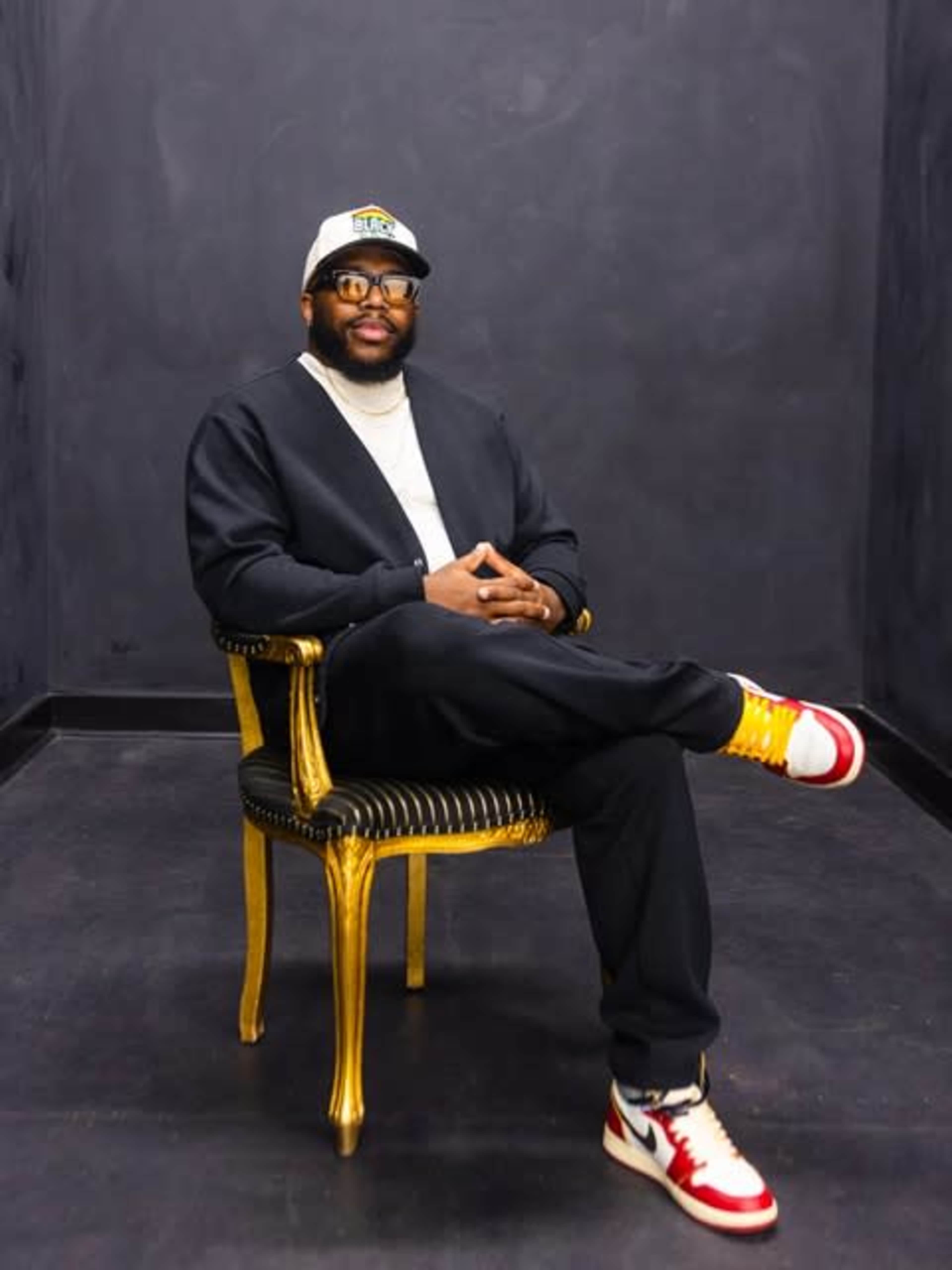 A man sits confidently on a gold-accented chair against a dark backdrop, wearing glasses and a baseball cap, with a relaxed posture and colorful sneakers.