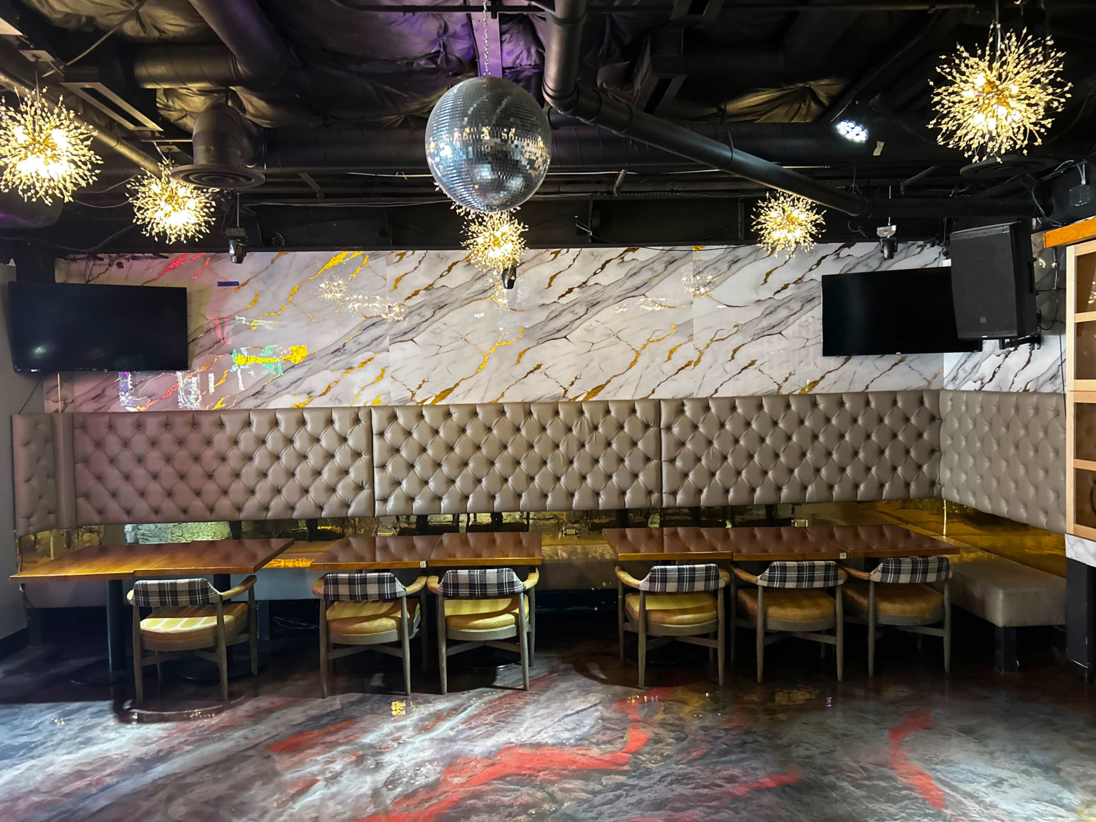 The image shows a modern restaurant interior with a long, tufted seating area, a glossy table, and decorative lighting fixtures, including a disco ball and two mounted televisions.