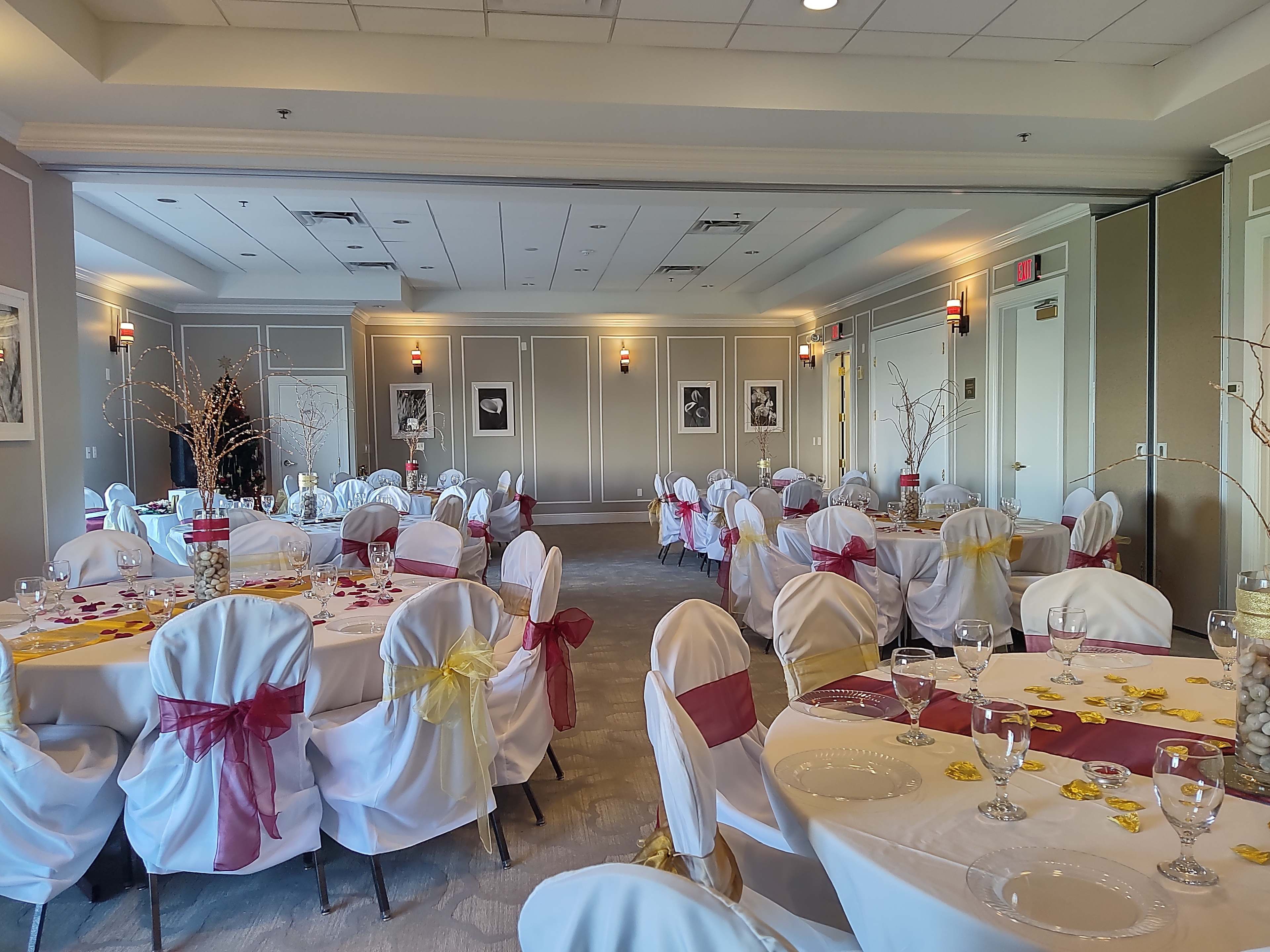 The image shows a banquet room arranged for an event, with round tables covered in white tablecloths and decorated with colored sashes, glassware, and scattered floral accents.