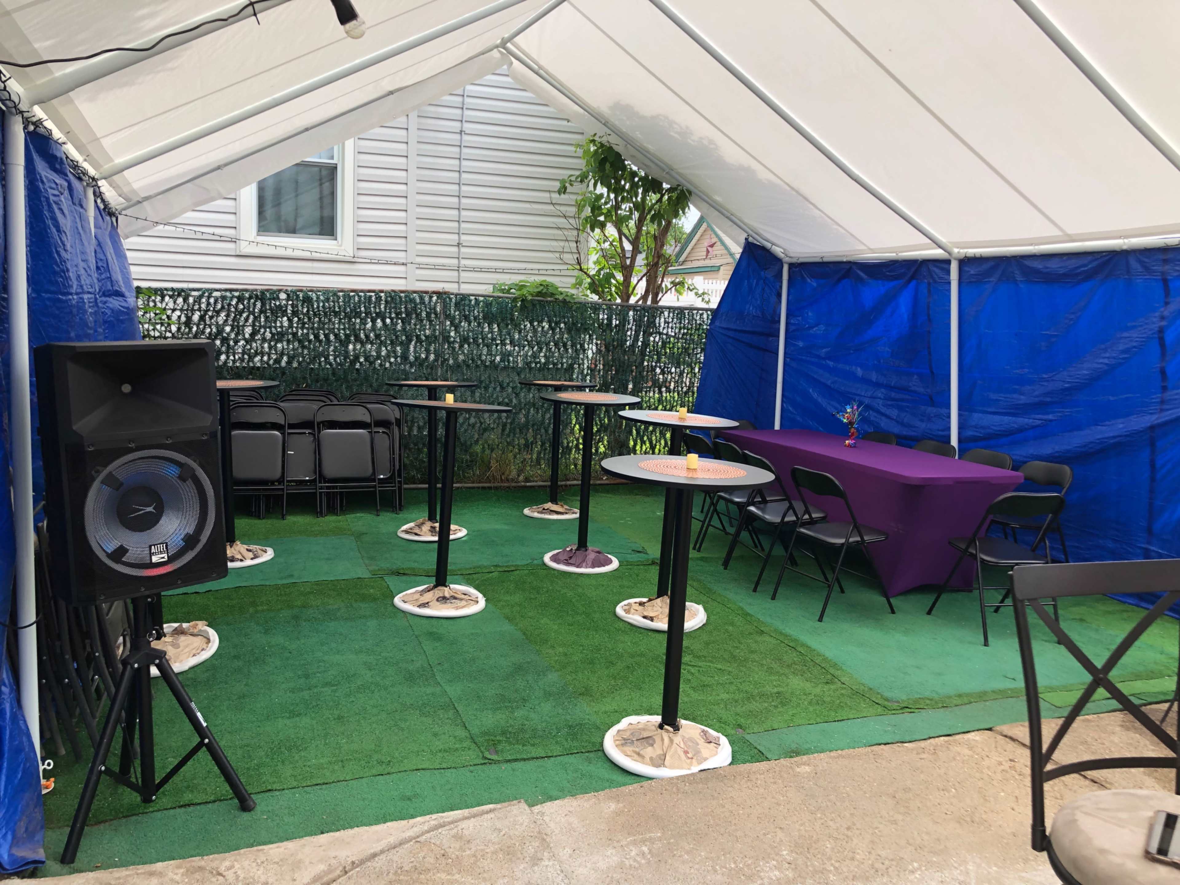 The image shows a tented area with several standing tables, chairs, and a purple tablecloth set up for a gathering.