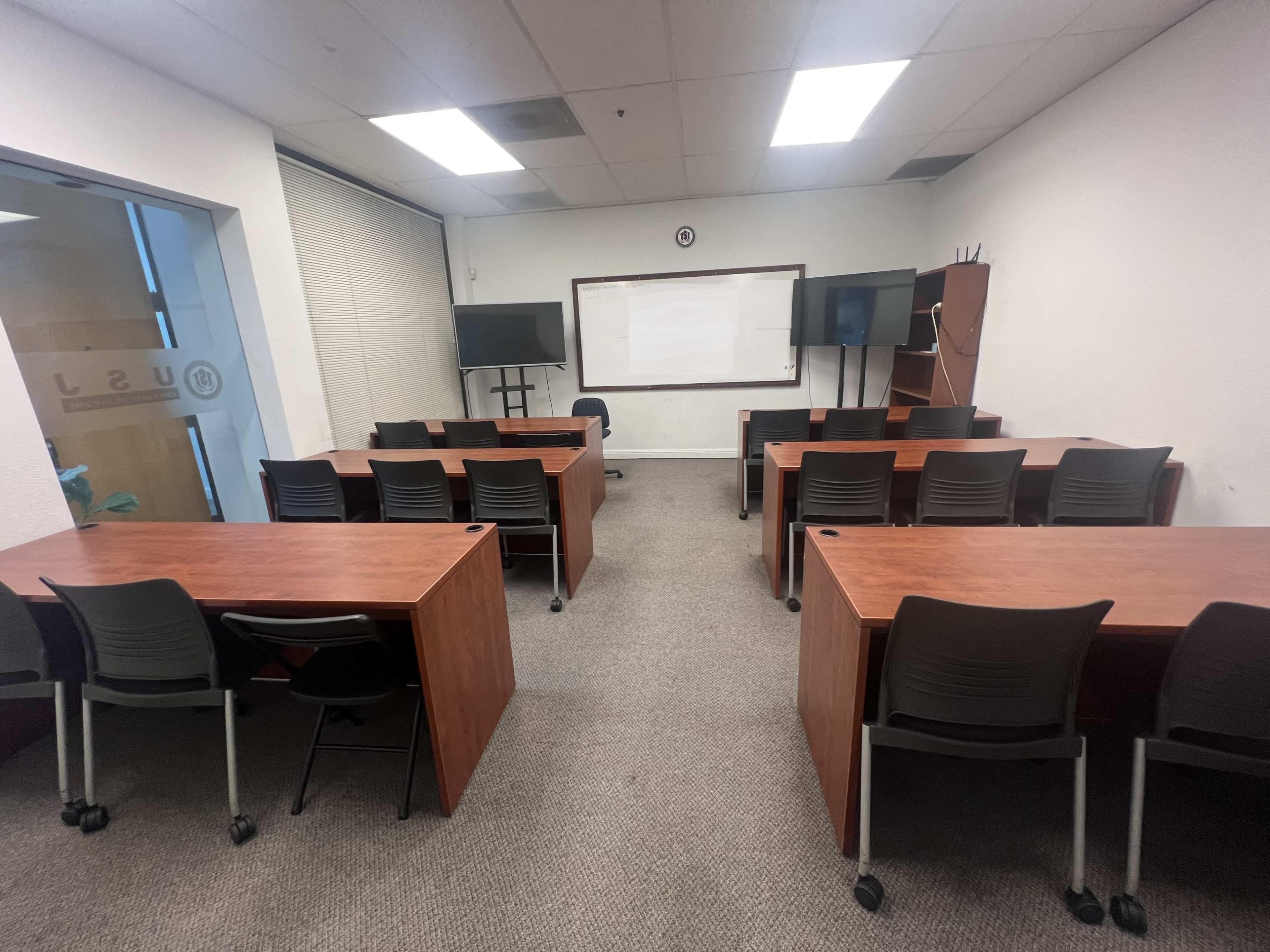 A classroom is arranged with several wooden tables and black chairs, facing a whiteboard and screens at the front.