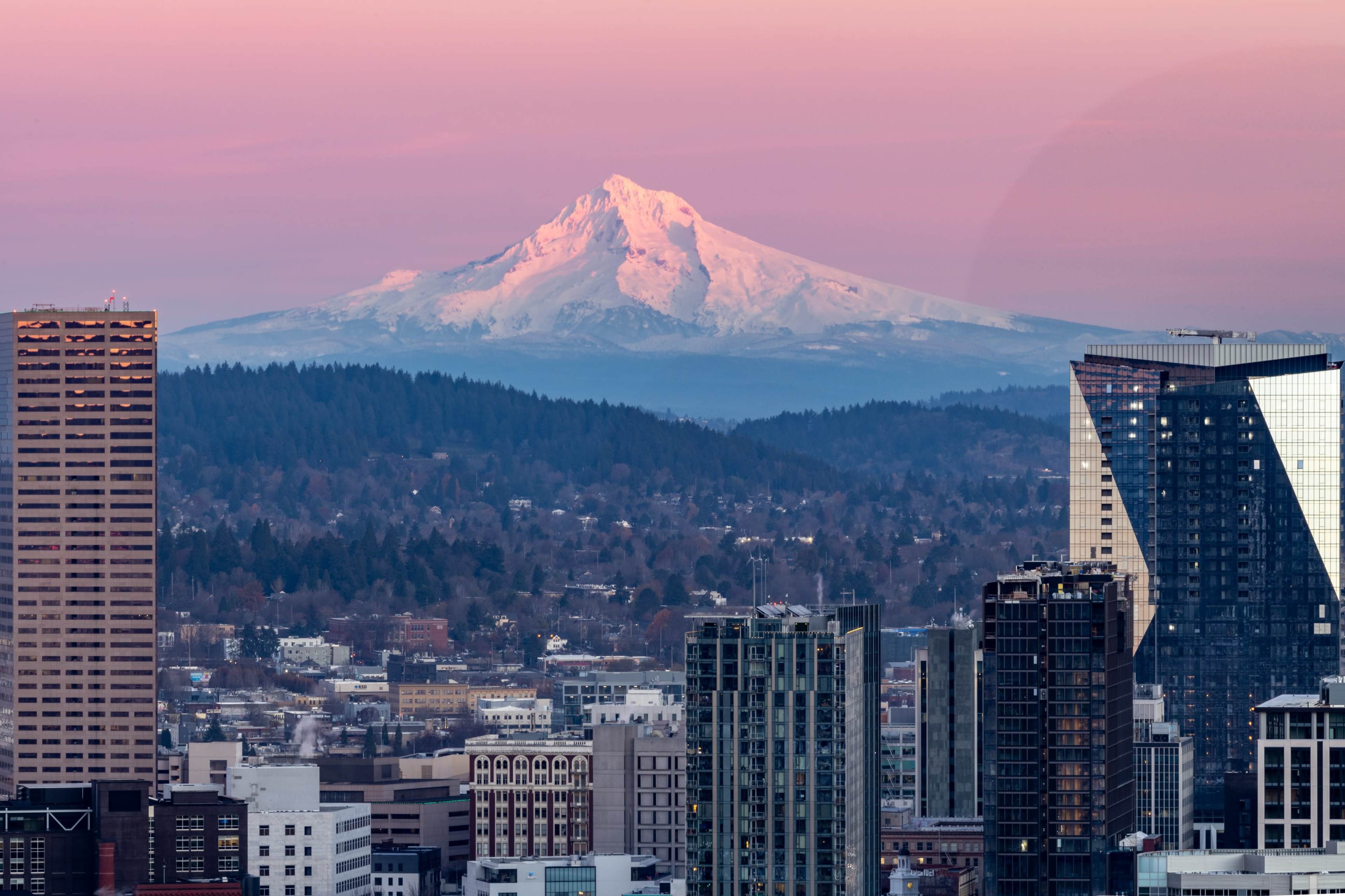 A snow-capped mountain rises in the background over a city skyline at dusk.