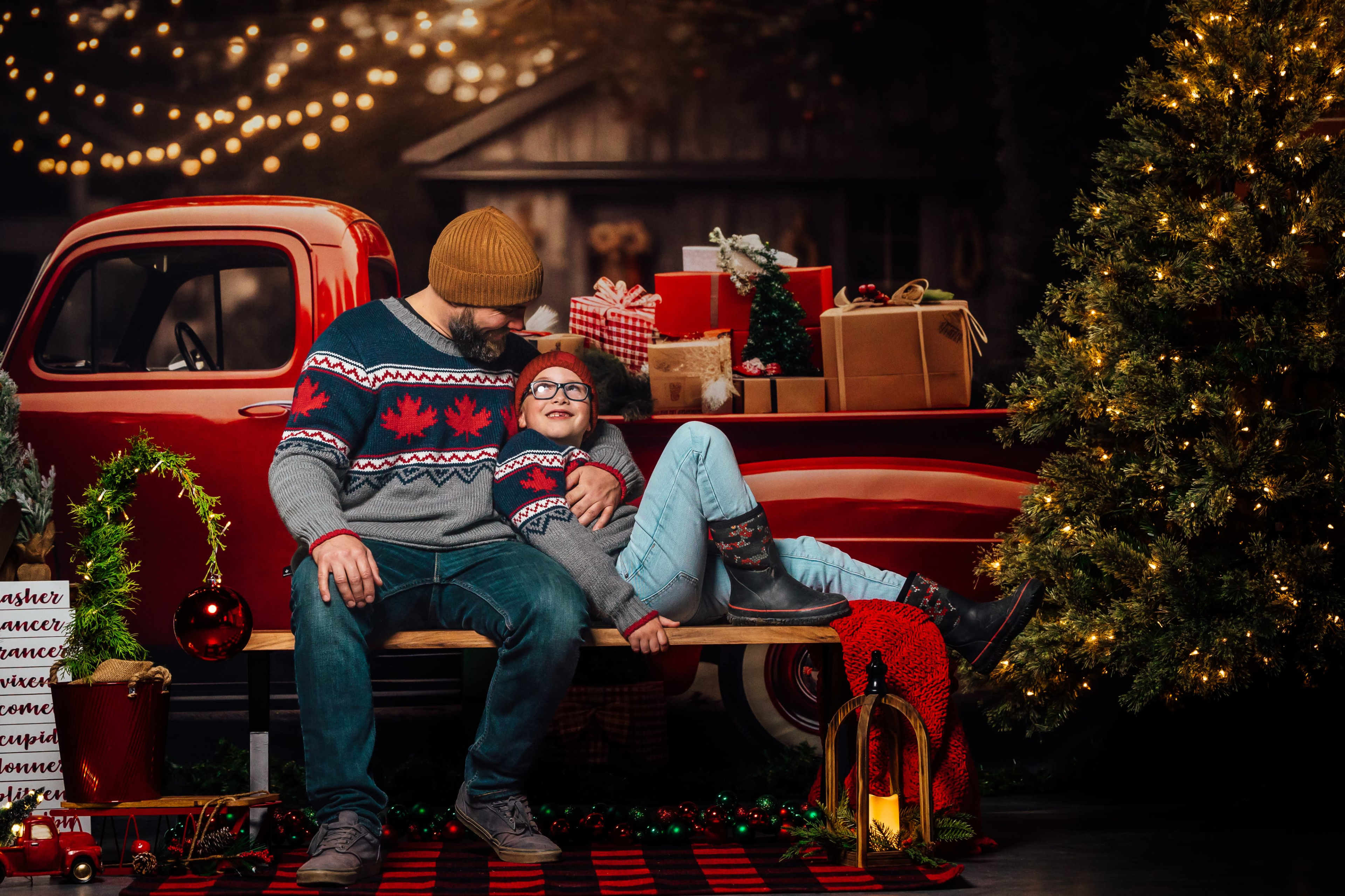 A man and a girl sit together on a bench in front of a vintage red truck decorated for Christmas, surrounded by presents and a brightly lit tree.