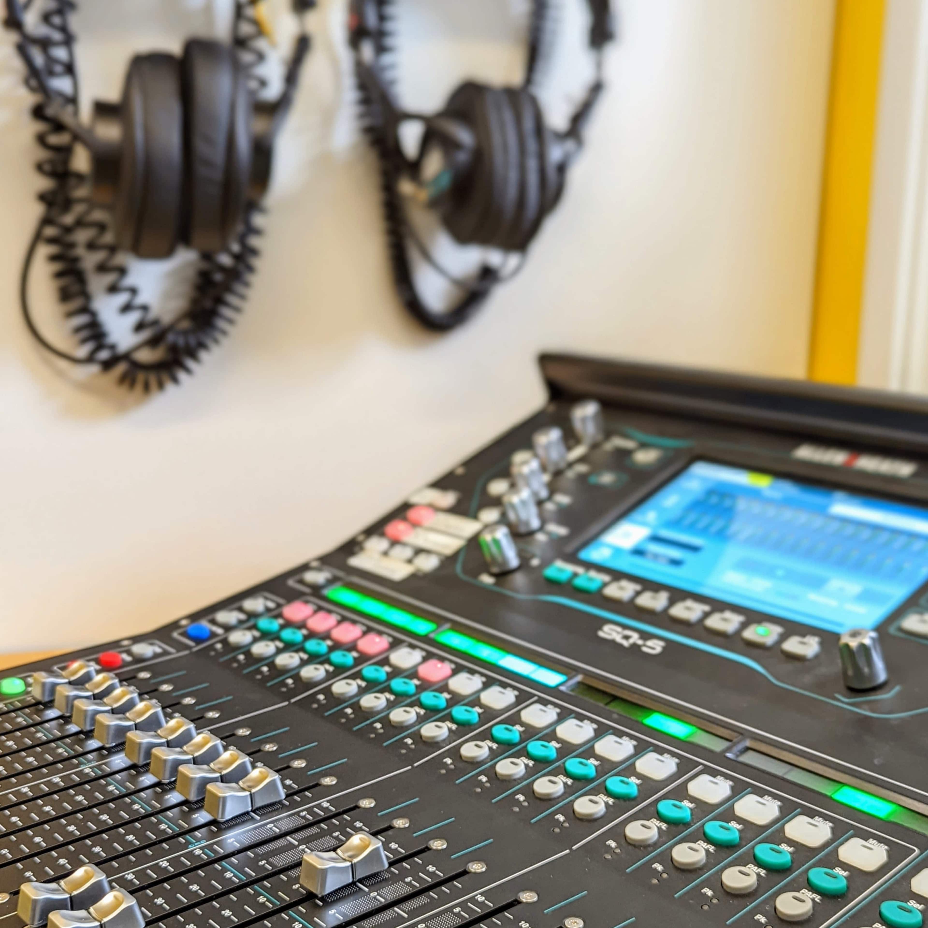 The image shows a digital mixing console with various knobs and sliders, accompanied by two pairs of headphones hanging on the wall.