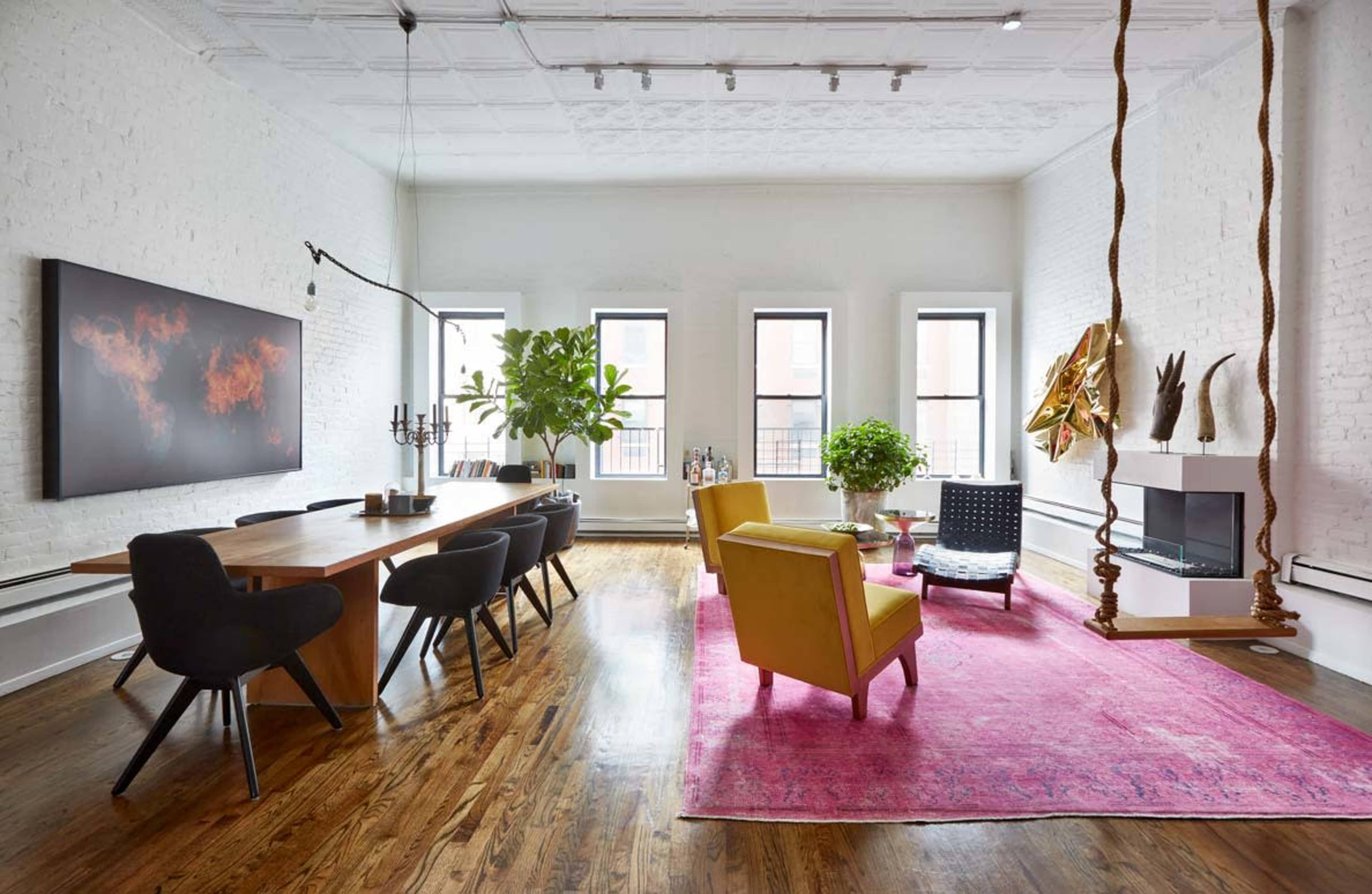 A bright living space featuring a wooden dining table, black dining chairs, a yellow armchair, a pink area rug, plants, and a modern fireplace.