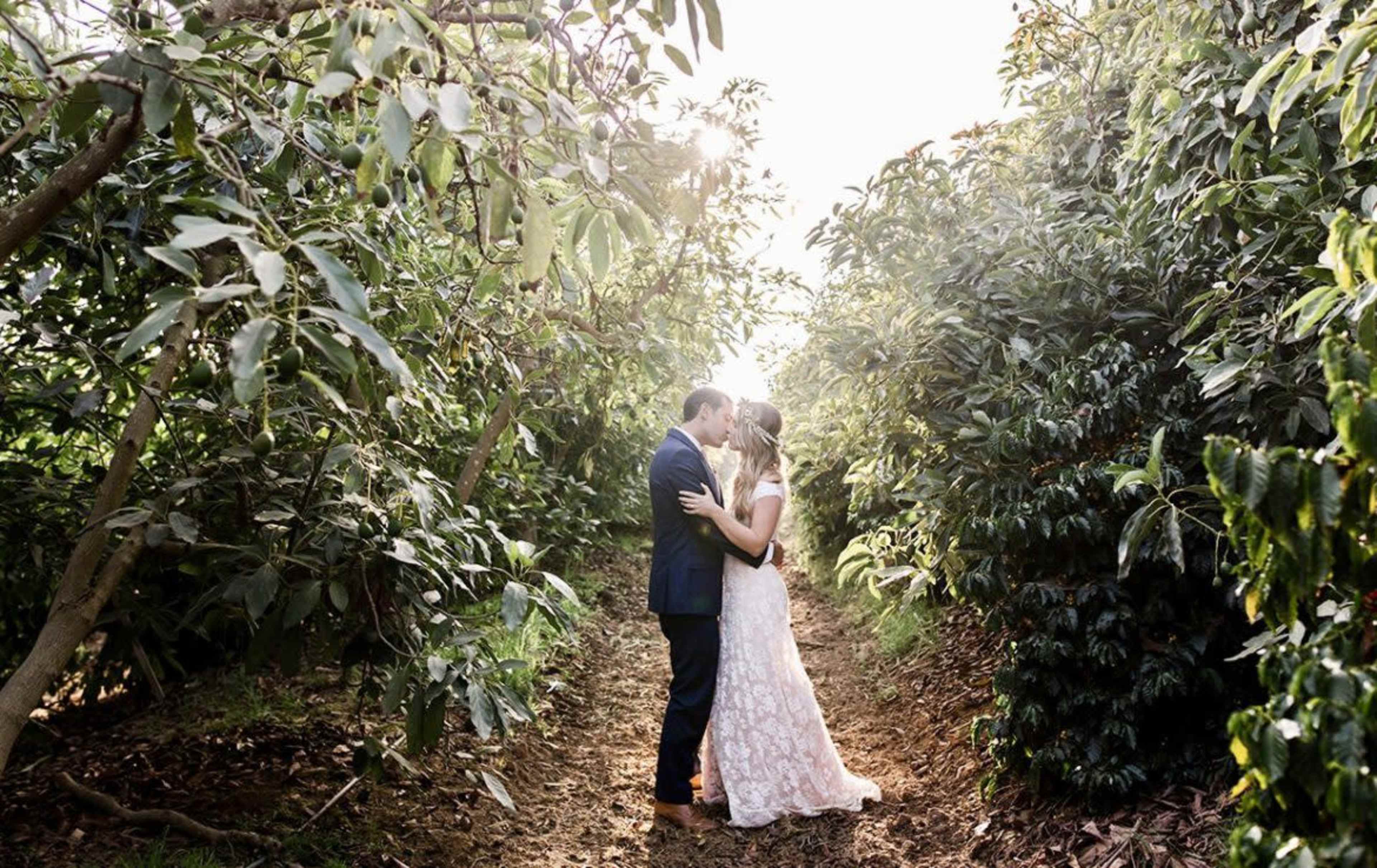 A couple stands in a lush, green pathway surrounded by trees, embracing and sharing a kiss.