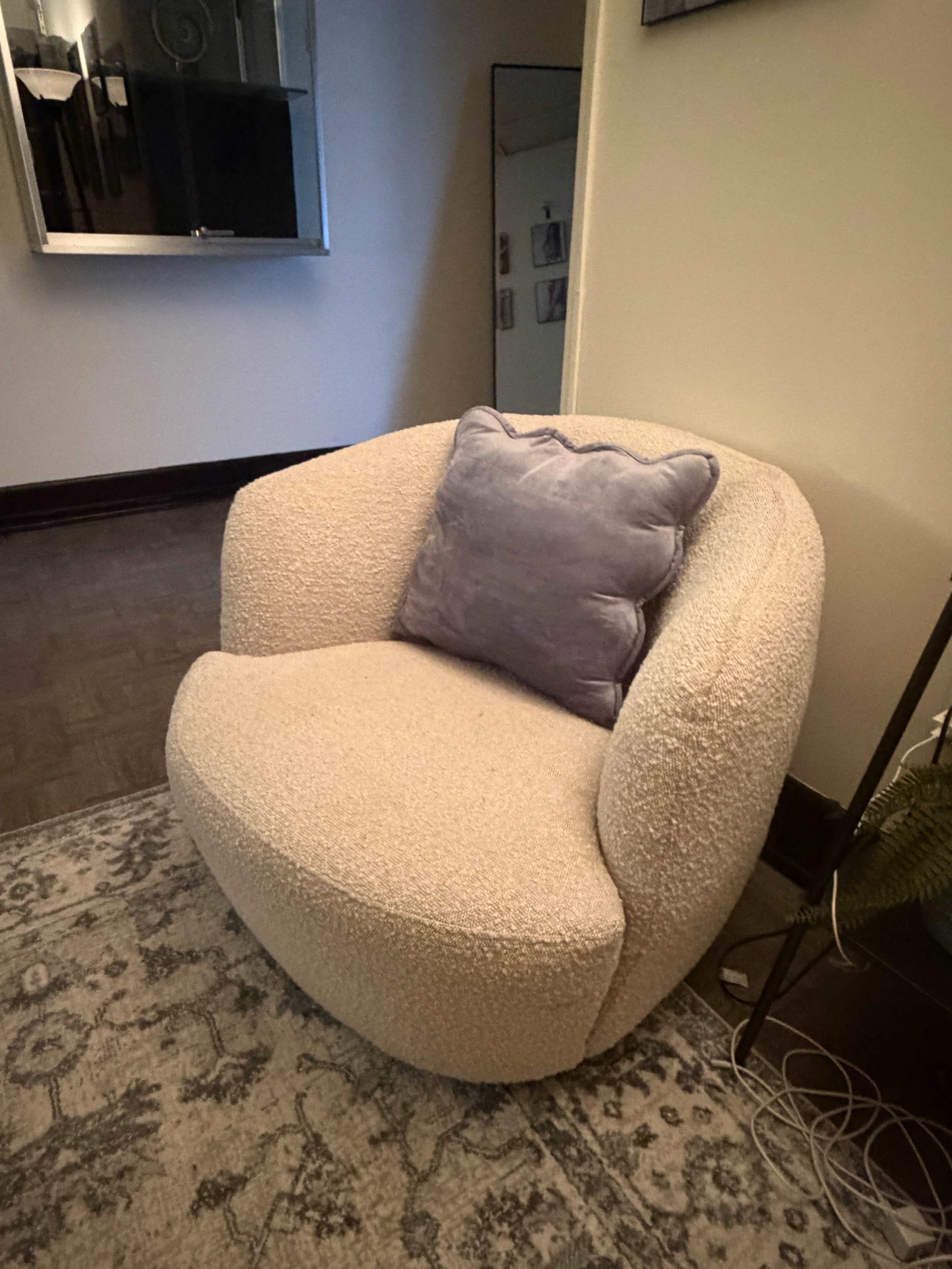 A light-colored, curved armchair with a plush pillow sits on a patterned rug in a corner of a room.