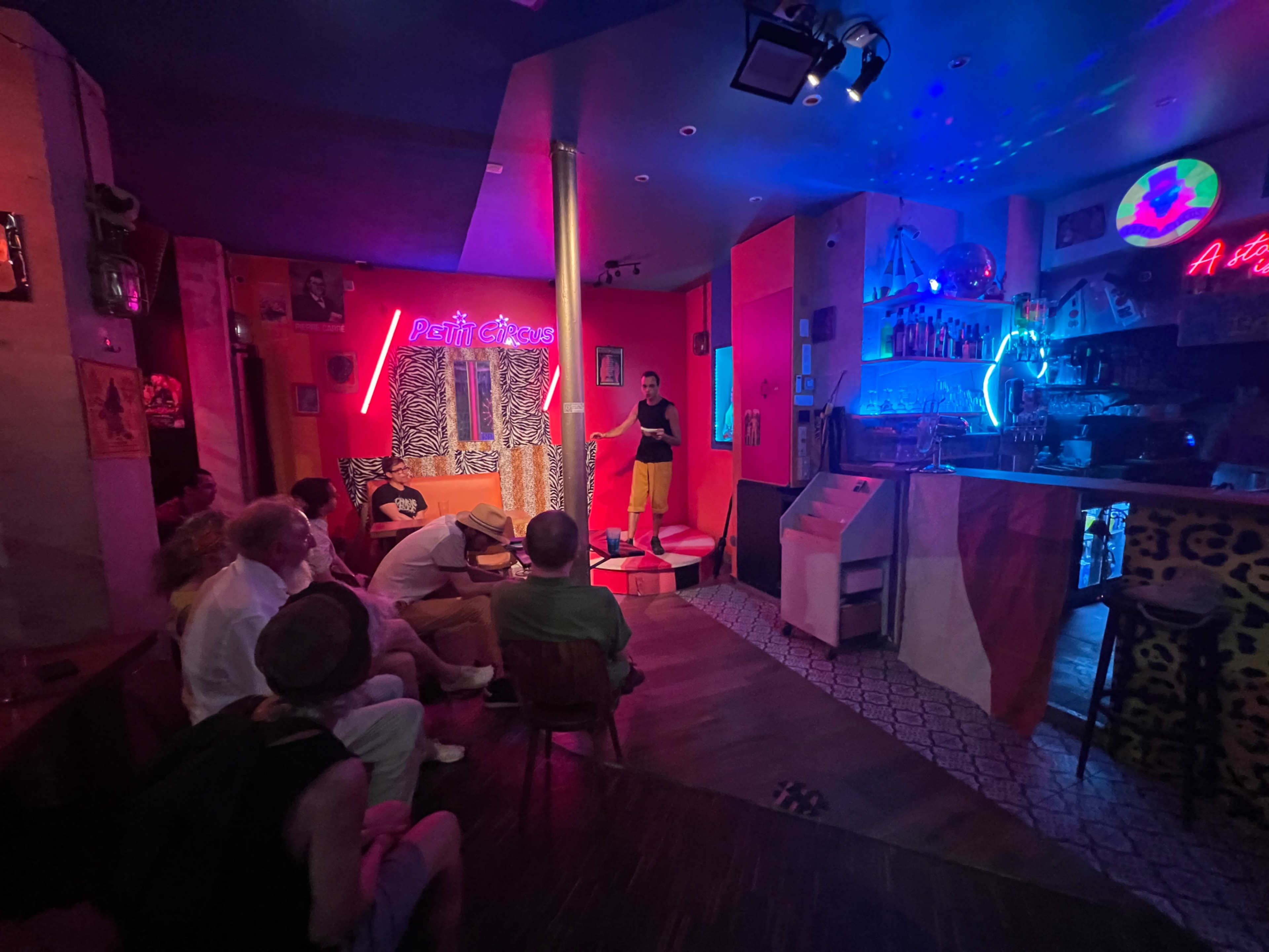 A performer stands on a small stage in a dimly lit bar, while an audience sits in chairs facing the stage.