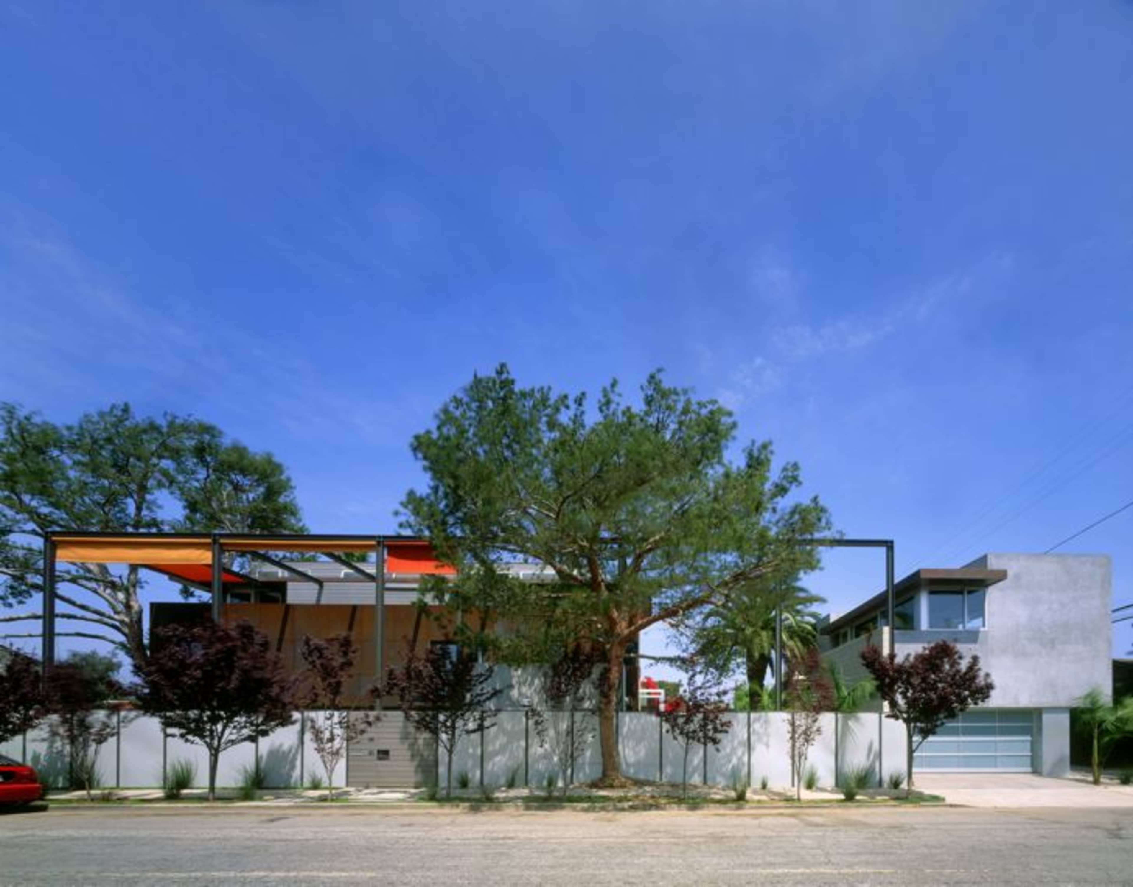 A modern residential building with a prominent tree in front is contrasted by a neighboring house featuring a garage, set against a clear blue sky.