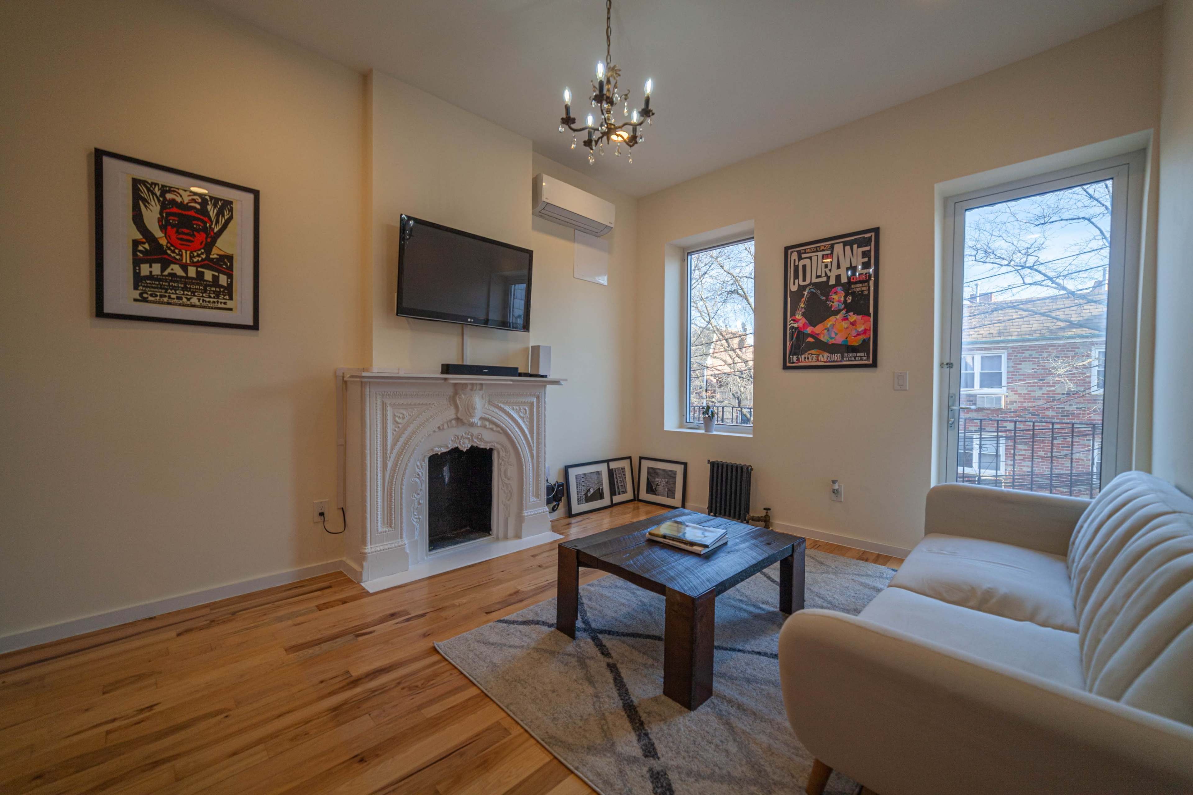 The living room features a chandelier, a white sofa, a wooden coffee table, and two windows with a view, along with decorative wall art and a fireplace.