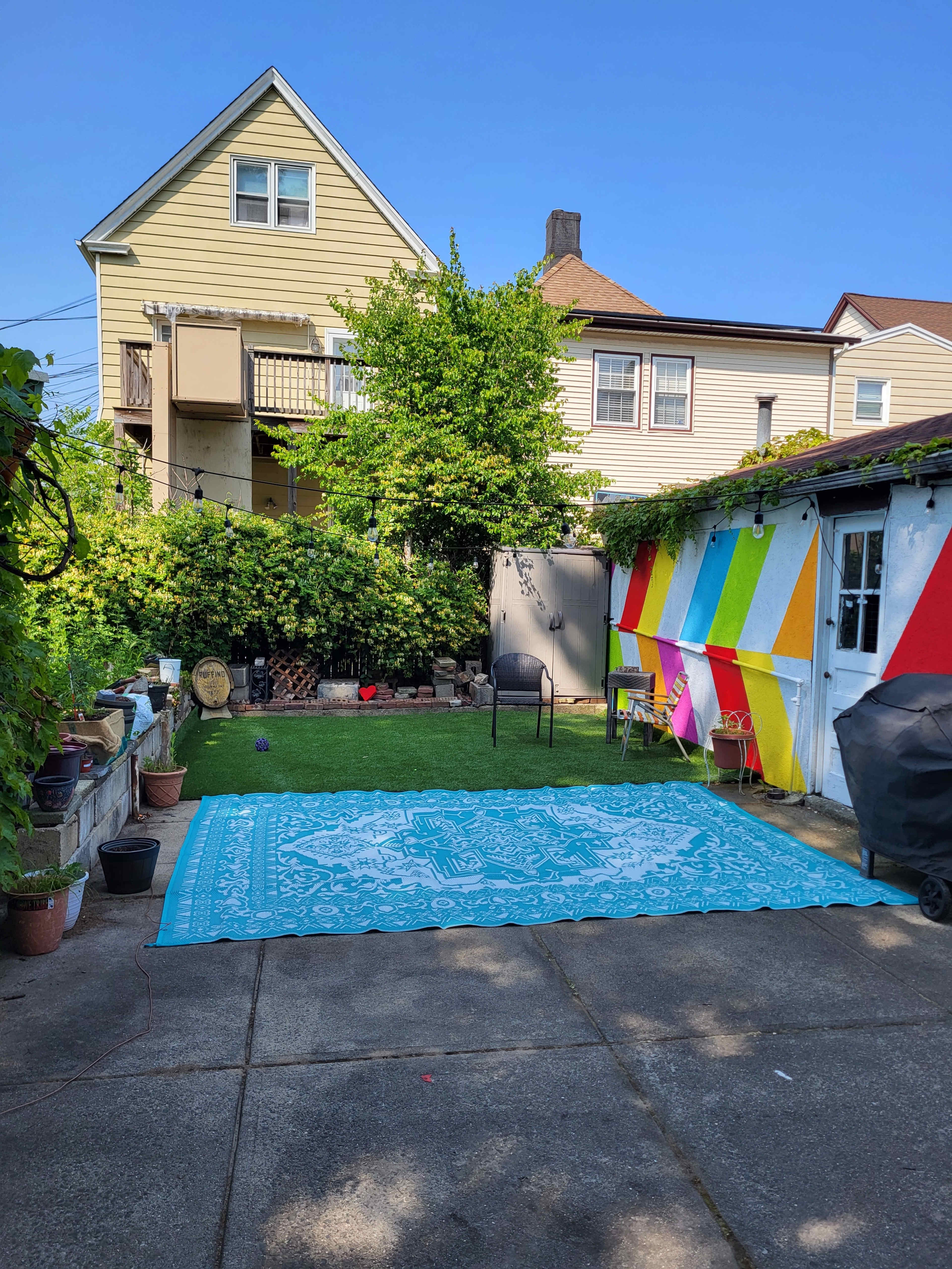 A colorful mural decorates one side of a backyard, which features a large patterned rug on the concrete, surrounded by potted plants and a chair.