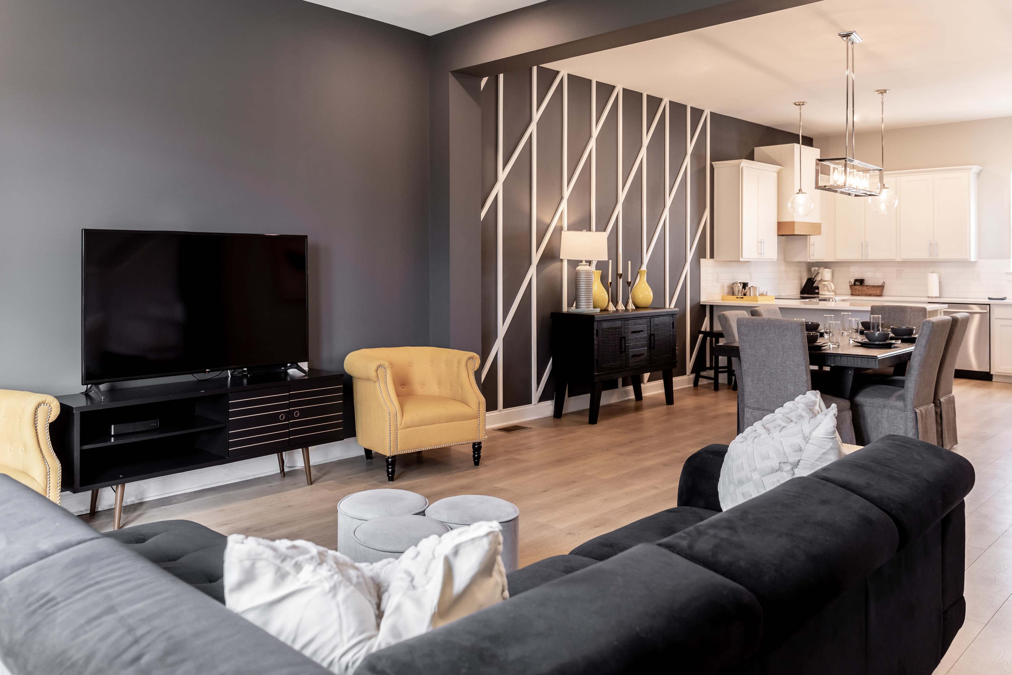 The image shows a modern living room with a dark gray wall, a large television, yellow accent chairs, a dining area, and a kitchen in the background.