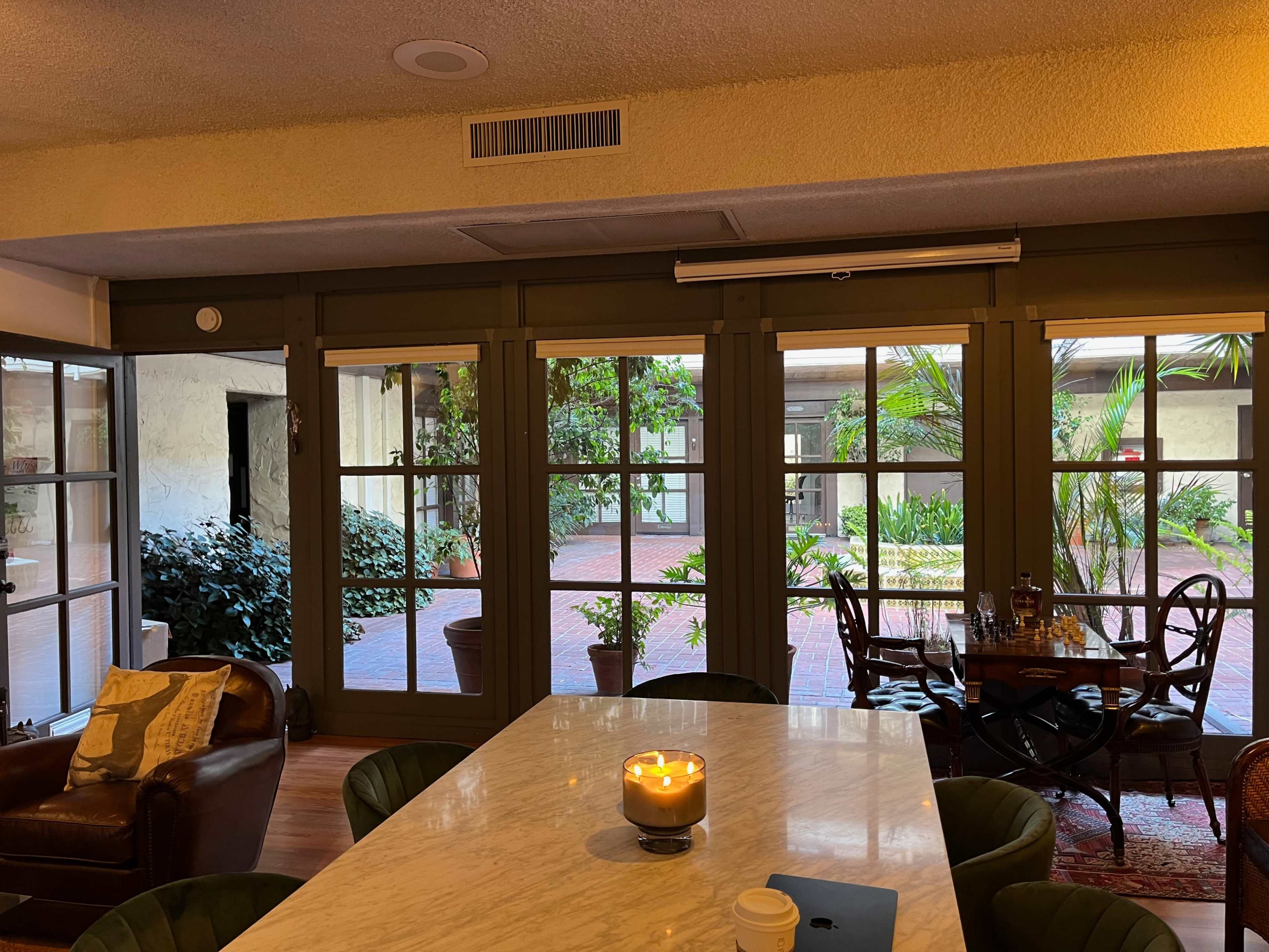A cozy indoor area with a marble table, a candle on it, and large windows overlooking a courtyard filled with greenery.
