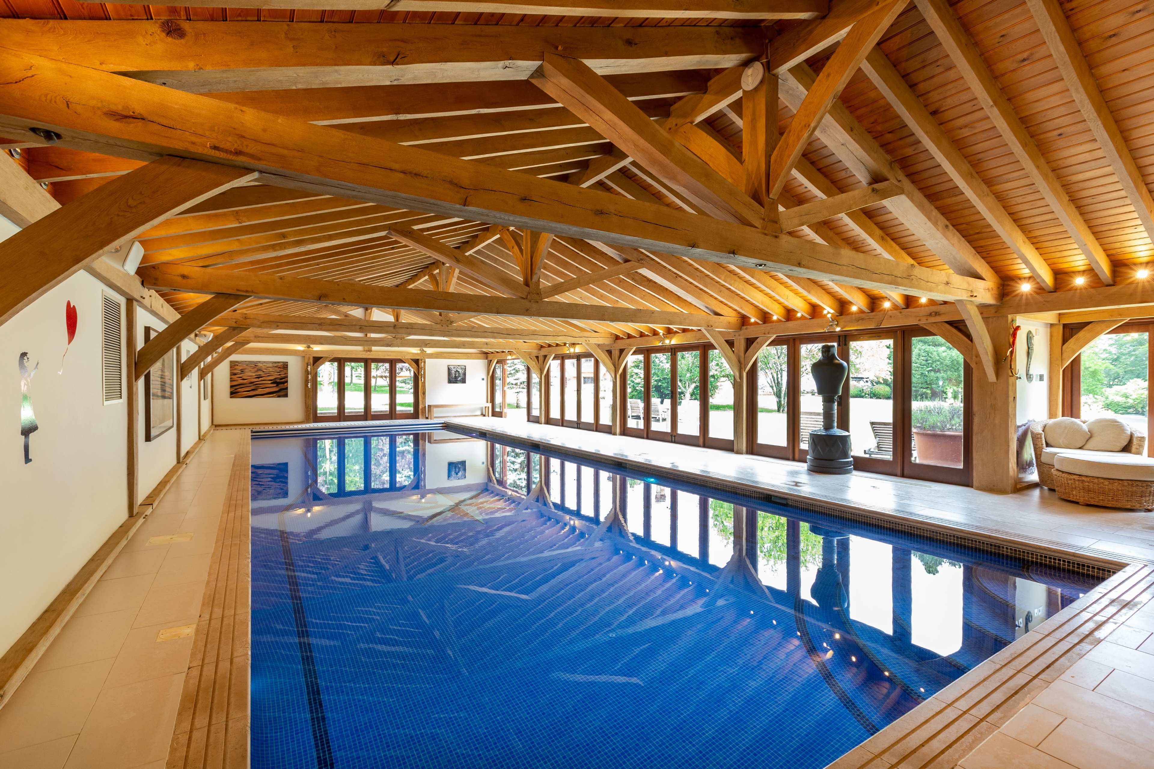 The image shows an indoor swimming pool with a wooden roof, large windows, and a serene ambiance.