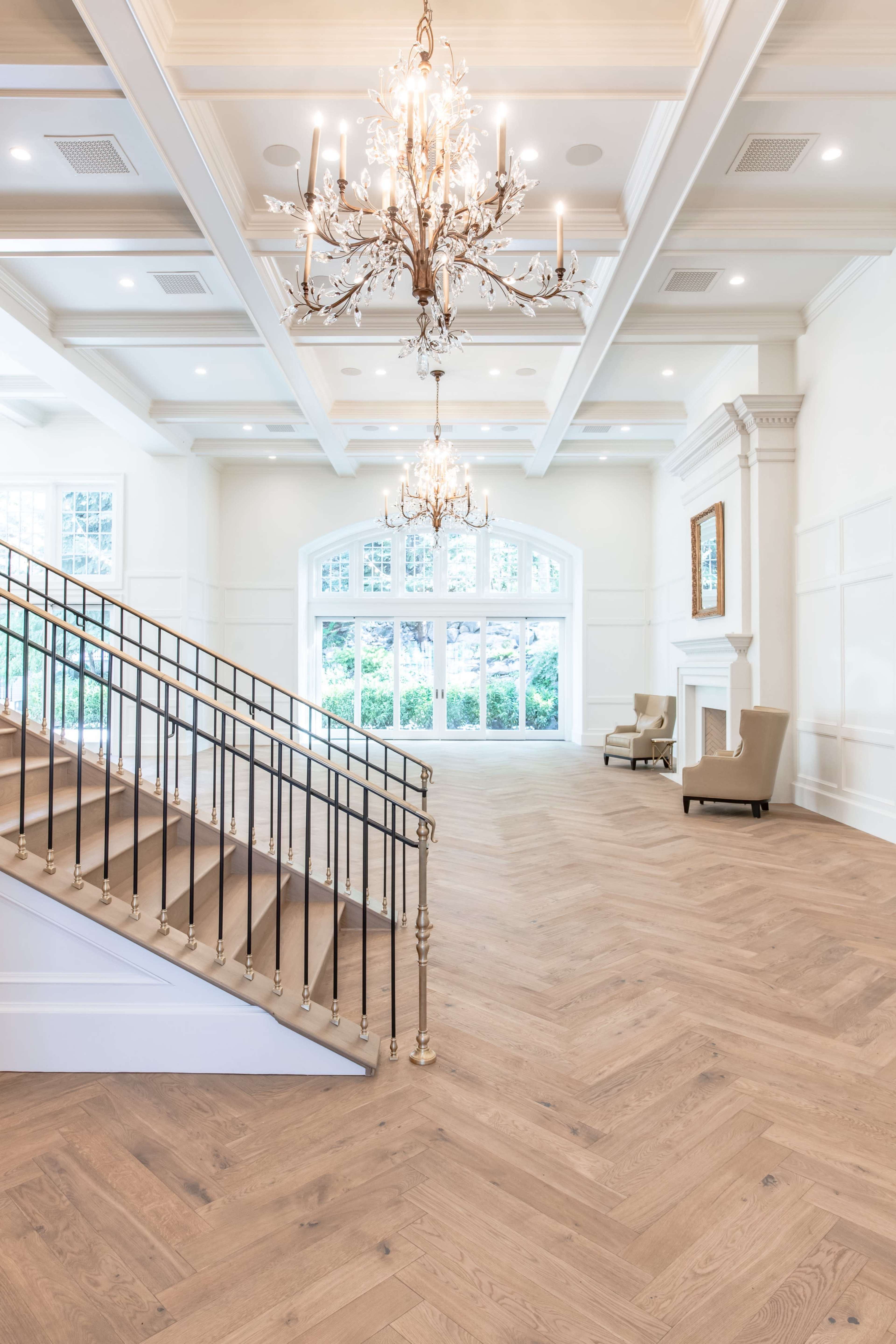 A spacious interior features a grand staircase with a wrought iron railing, elegant chandeliers, and large windows overlooking a garden.