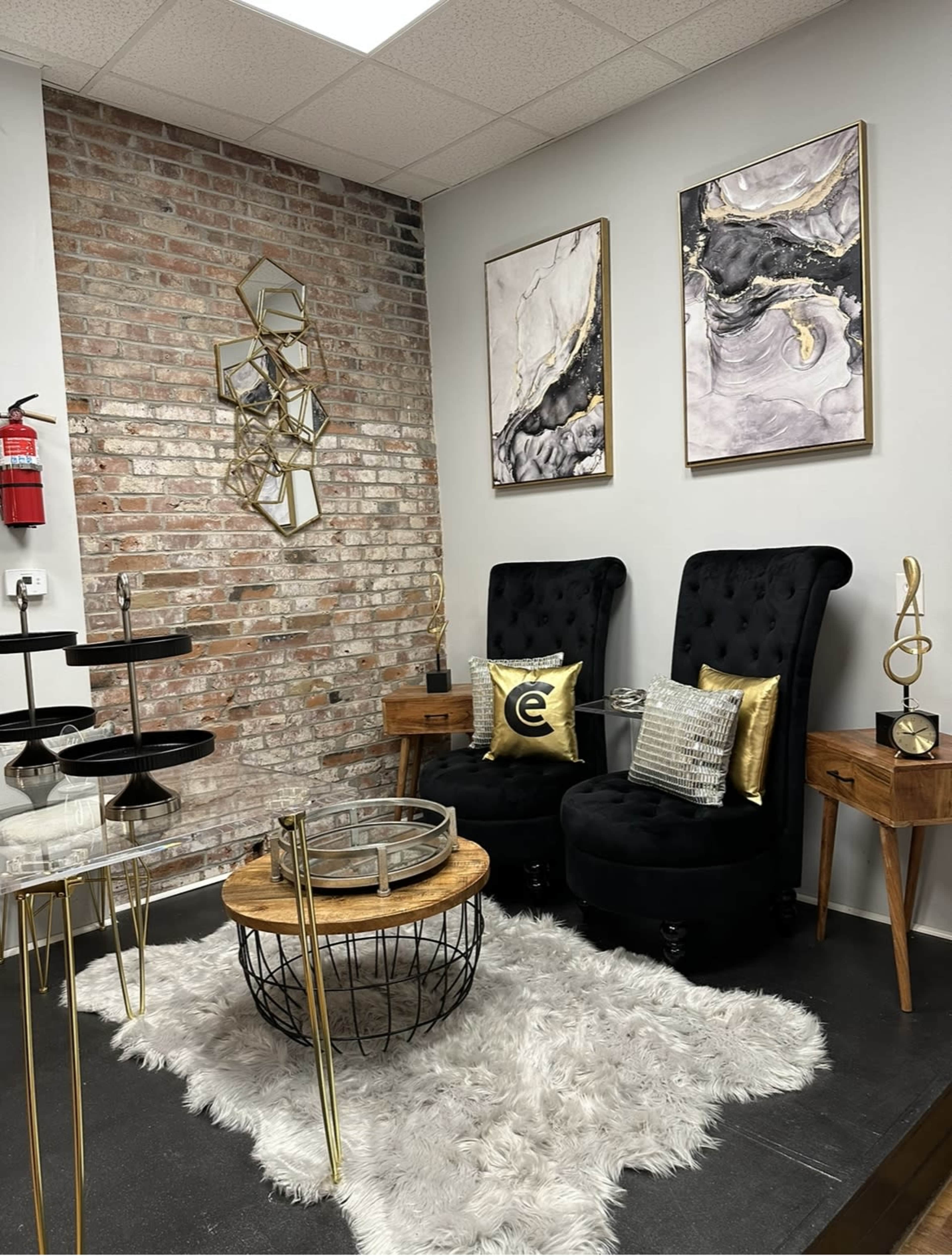 The scene features a stylish seating area with two black tufted chairs, a round coffee table on a plush white rug, decorative wall art, and a modern mirror, all set against a brick wall.