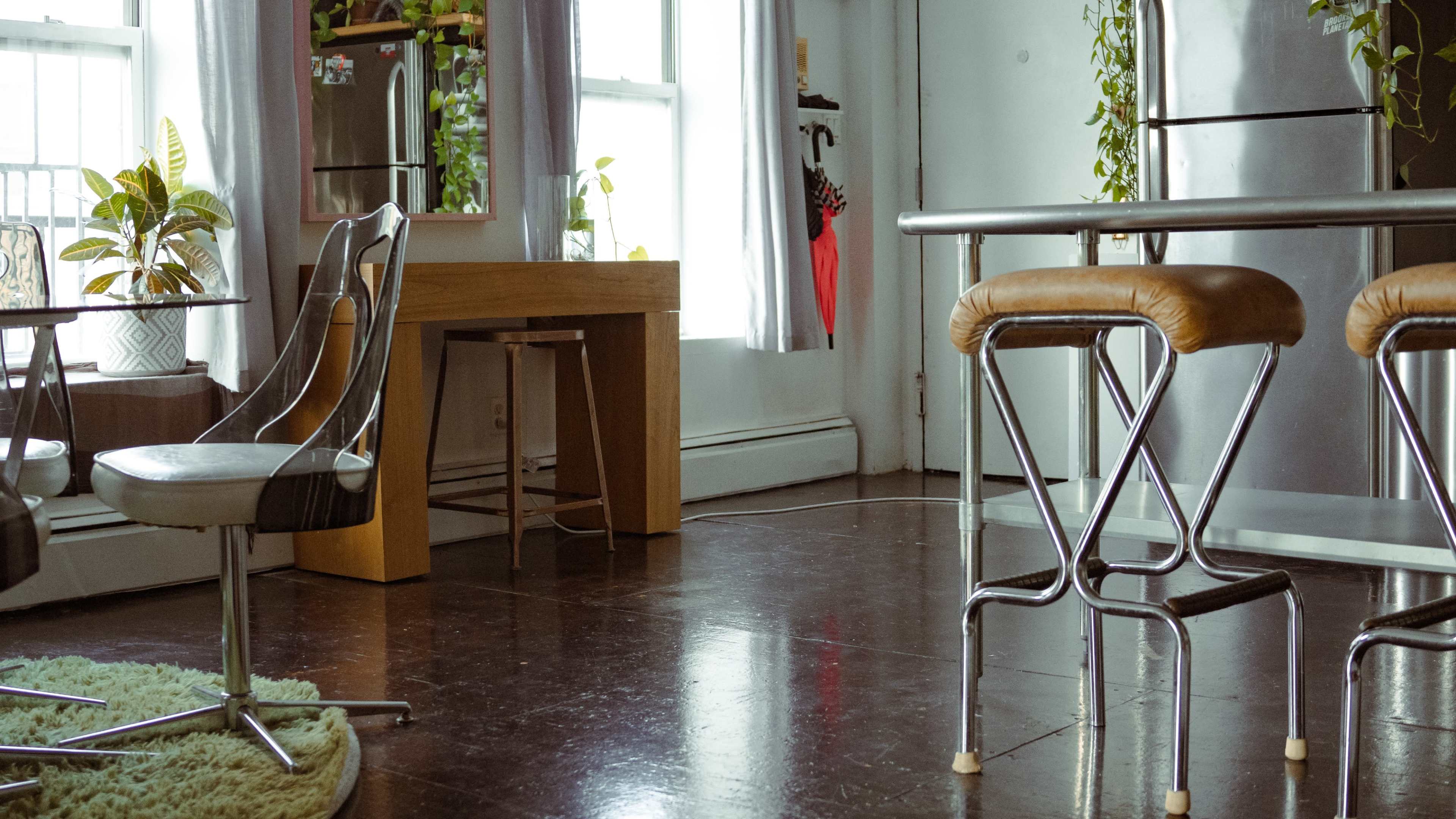 The scene features a modern kitchen area with two metal bar stools and a wooden table, complemented by houseplants and large windows letting in natural light.