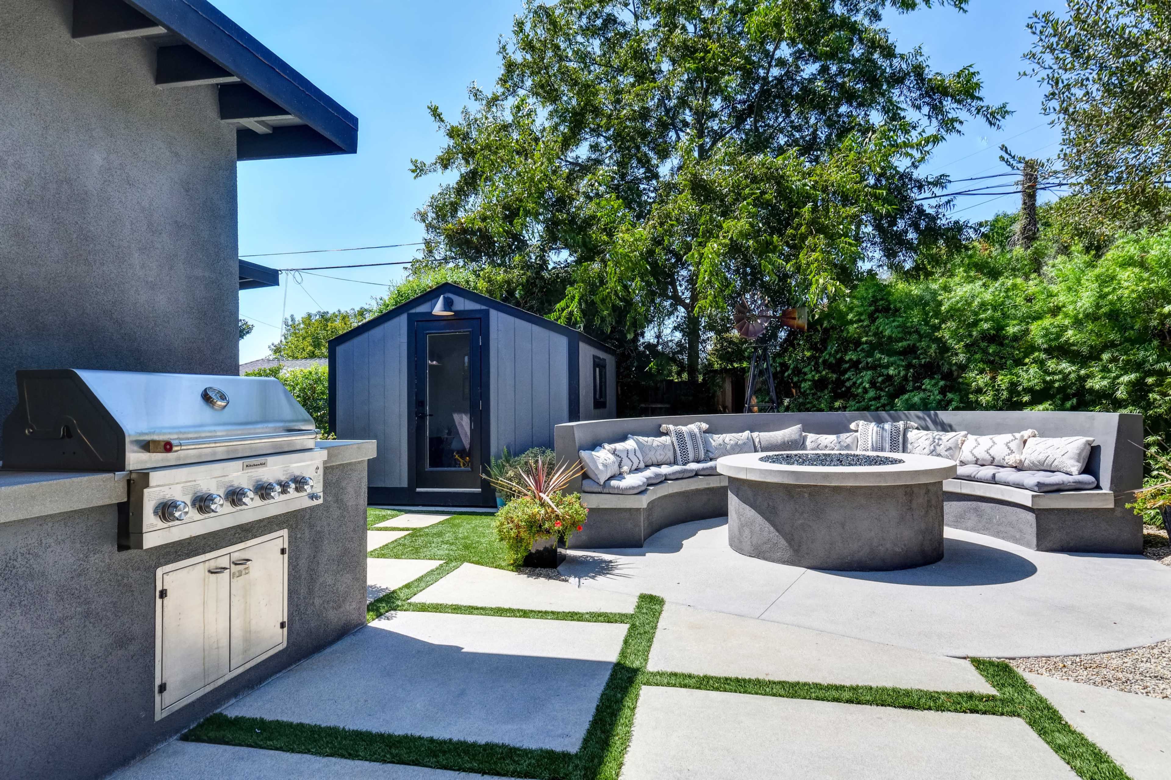 A modern outdoor space features a stainless steel grill, a circular fire pit surrounded by a curved seating area, and a small shed, all set against a backdrop of greenery.
