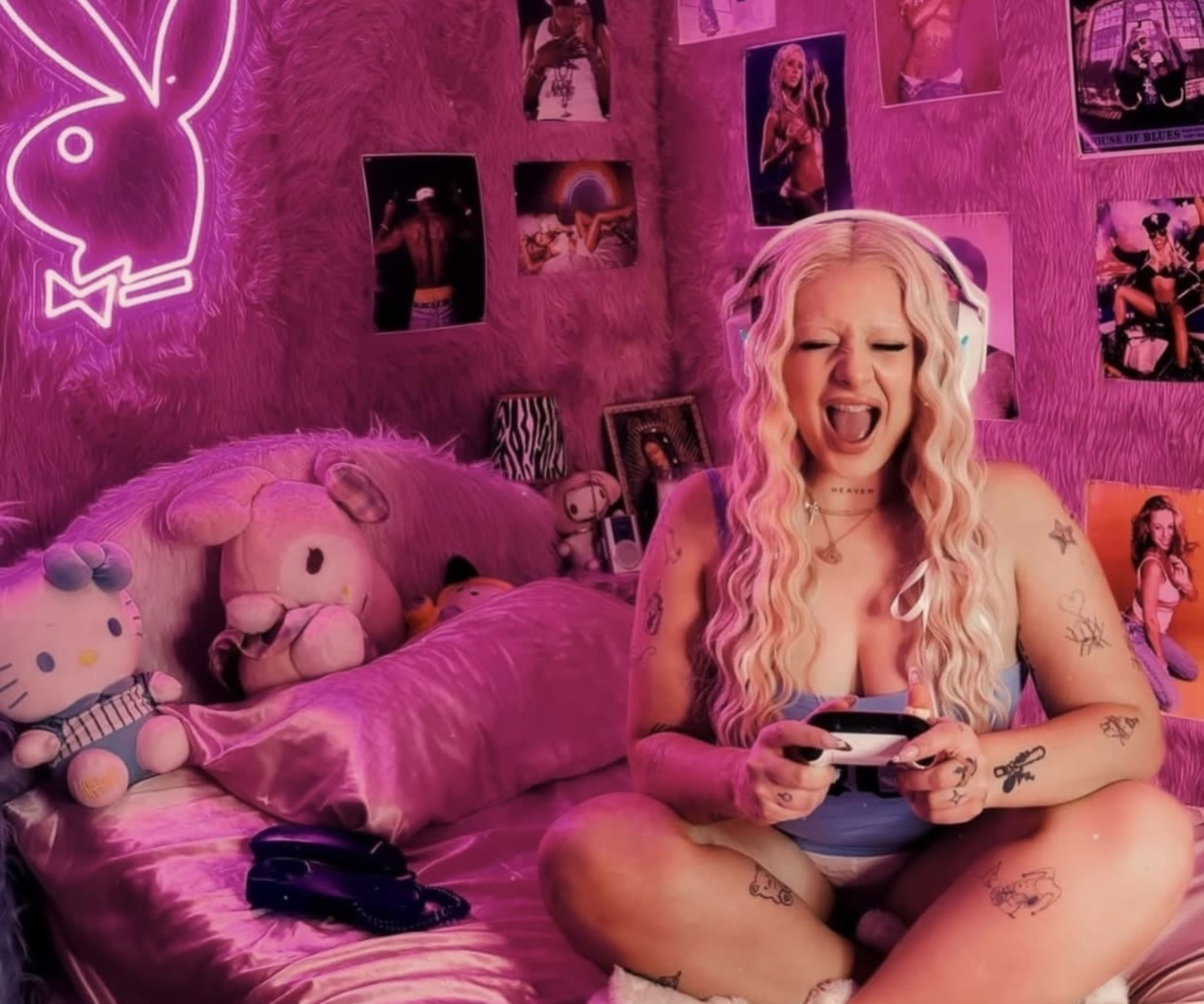 A person sits on a bed surrounded by plush toys and posters, holding a gaming controller in a room illuminated with pink lighting.