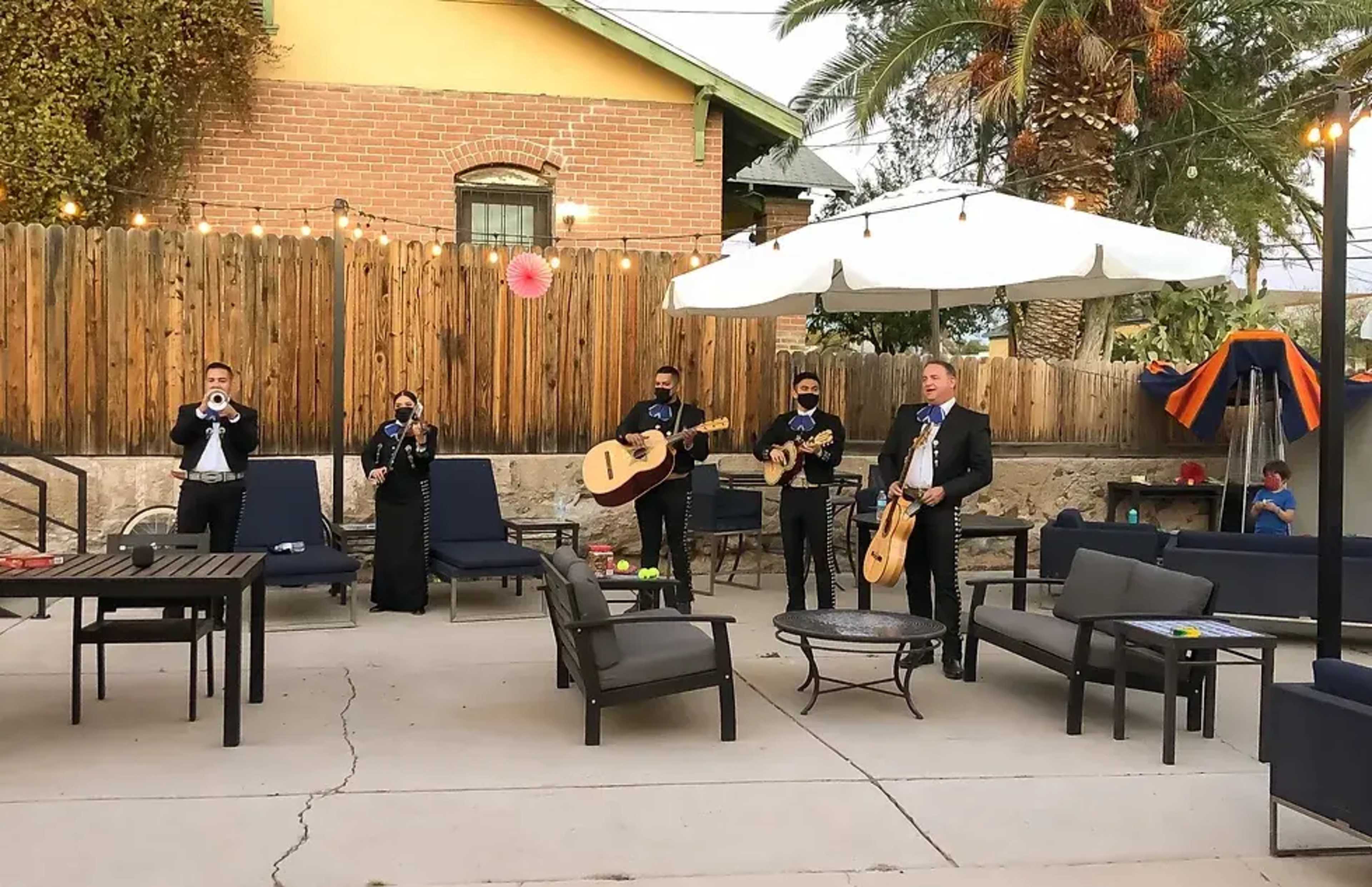 A group of musicians dressed in formal attire is performing with various instruments in a patio area surrounded by lounge furniture and string lights.