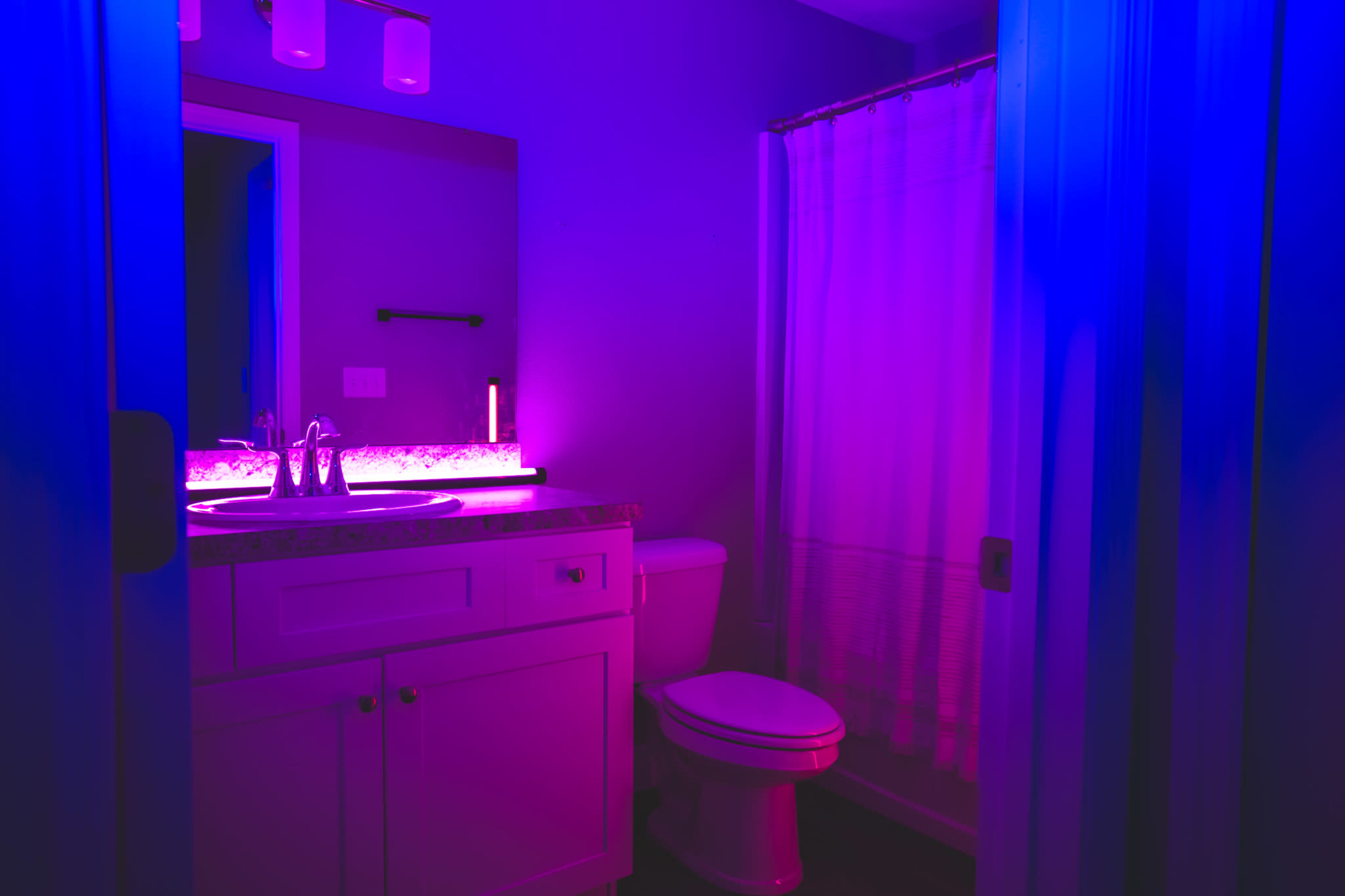 A bathroom features a sink with a lit mirror, a toilet, and a shower curtain, illuminated by purple and blue lighting.