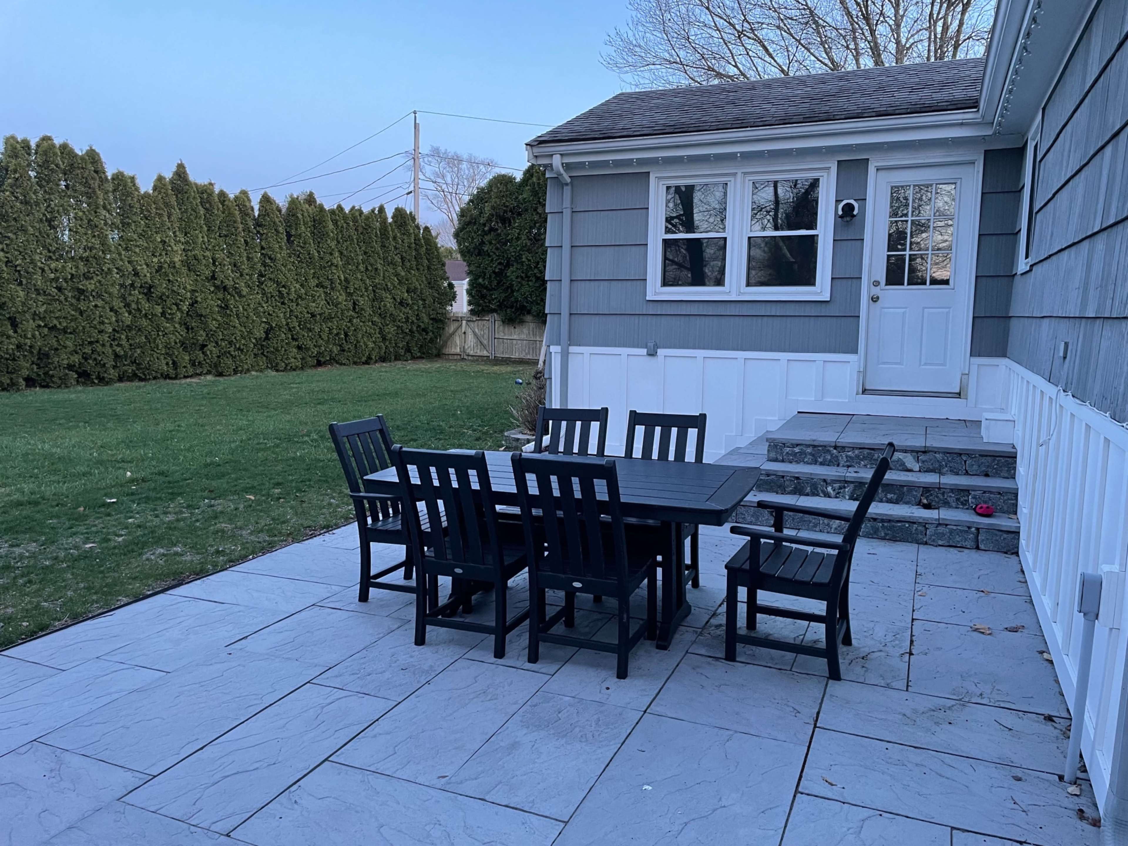 A black dining table with several matching chairs is set on a stone patio next to a gray house and a neatly trimmed lawn.