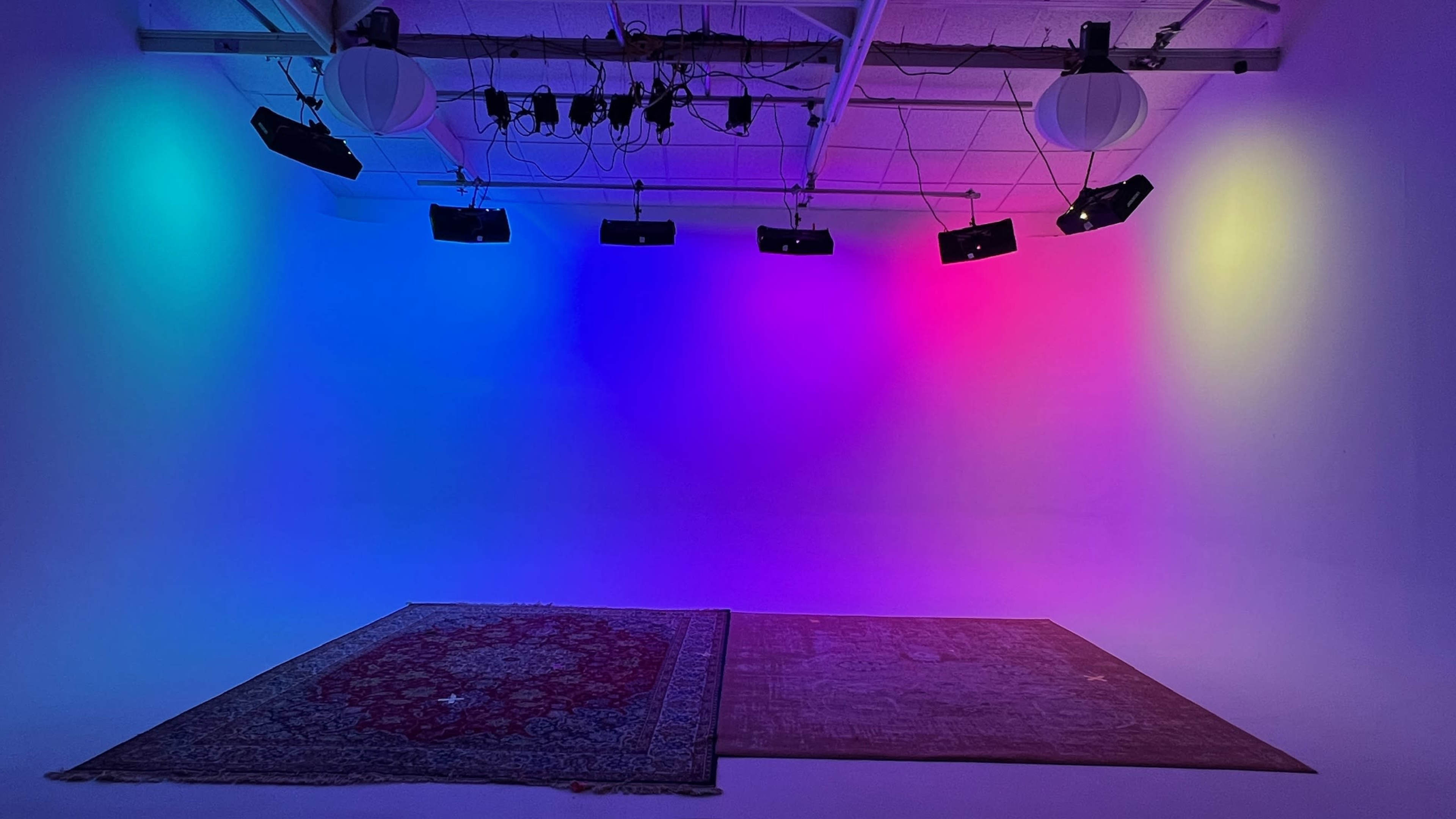 The image shows a brightly lit studio space with colored lights casting a gradient on the walls and two area rugs placed on the floor.