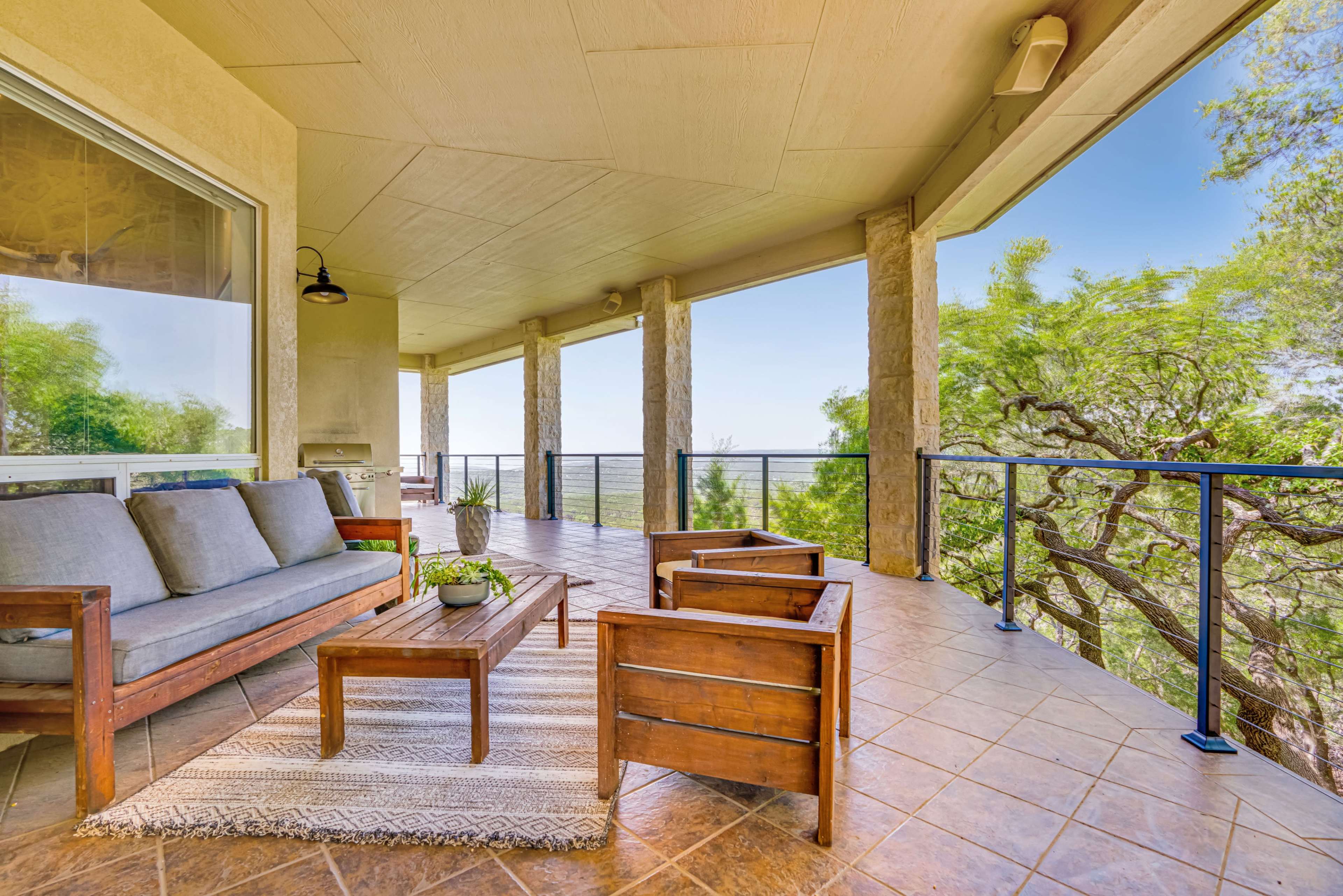 A covered porch features a gray sofa, wooden chairs, and a table surrounded by greenery and a panoramic view.