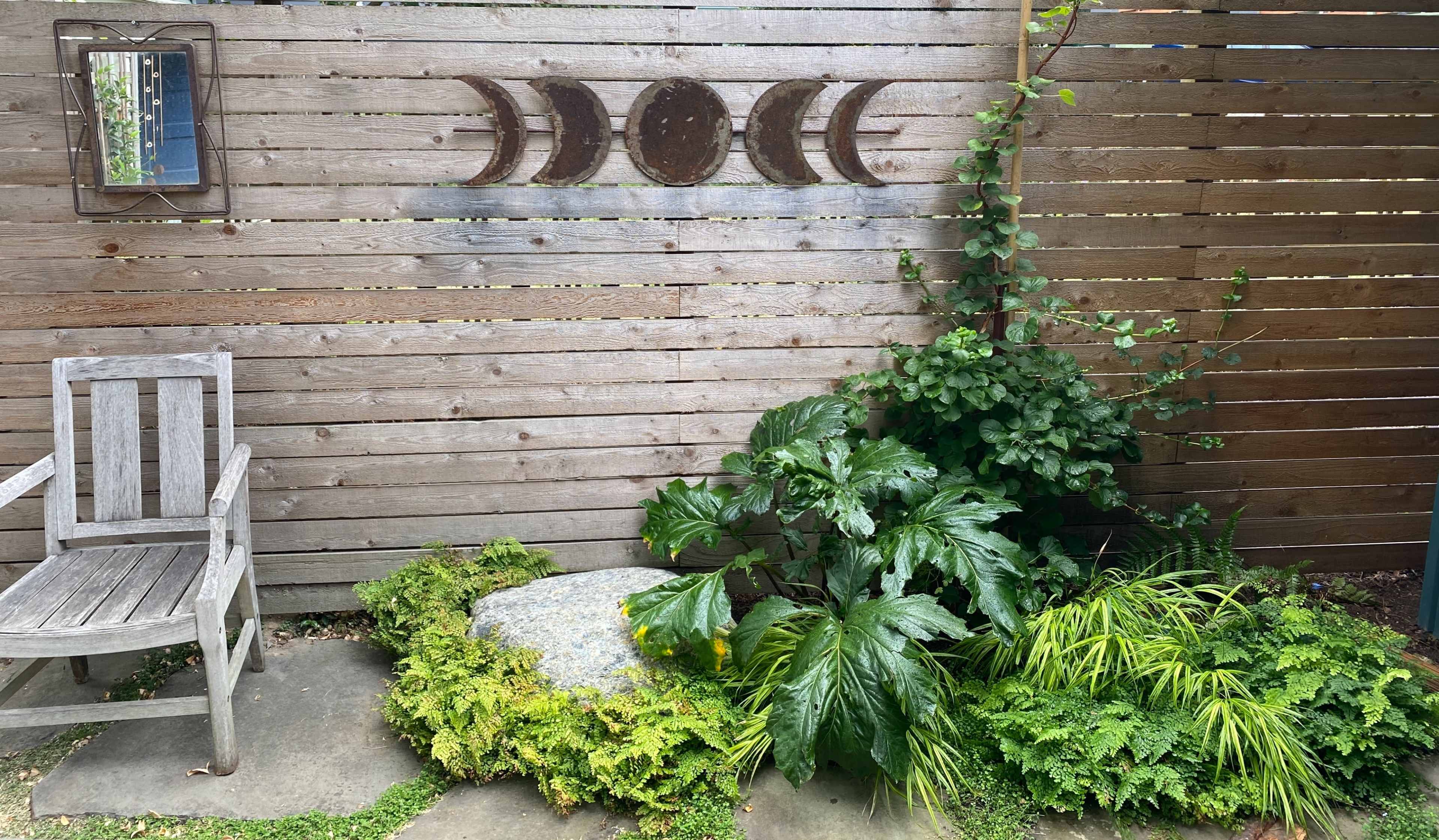 A wooden garden bench sits beside a stone surrounded by various green plants against a wooden fence featuring crescent moon decorations.