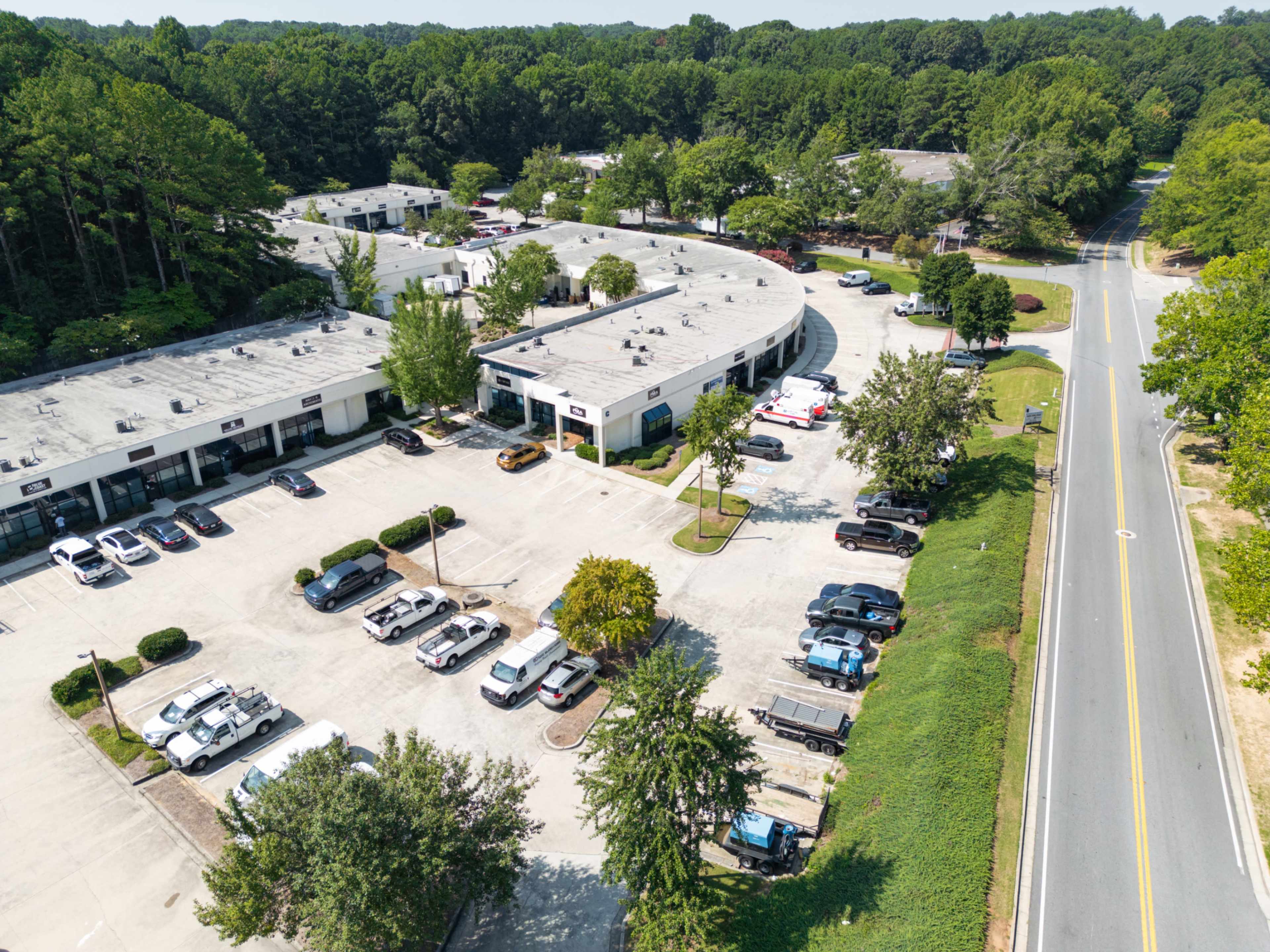 An aerial view shows a commercial area with several single-story buildings and a parking lot filled with vehicles, surrounded by trees and green space.